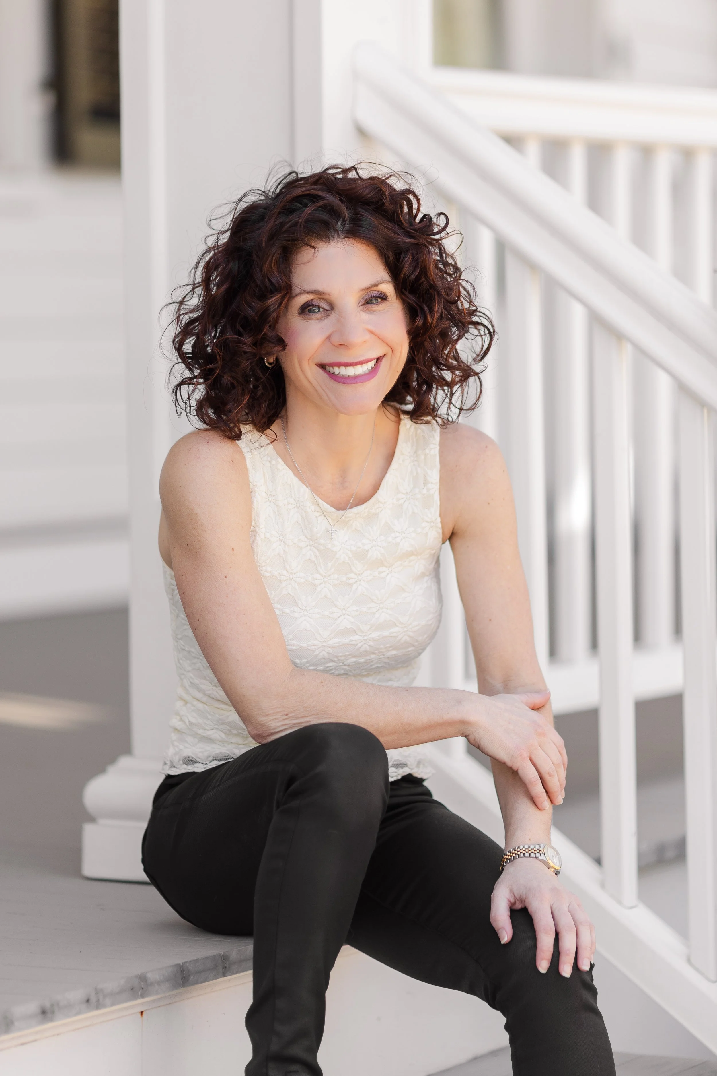 A woman with curly brown hair sitting on white stairs outside, smiling and looking at the camera, dressed in a sleeveless white lace top and black pants.