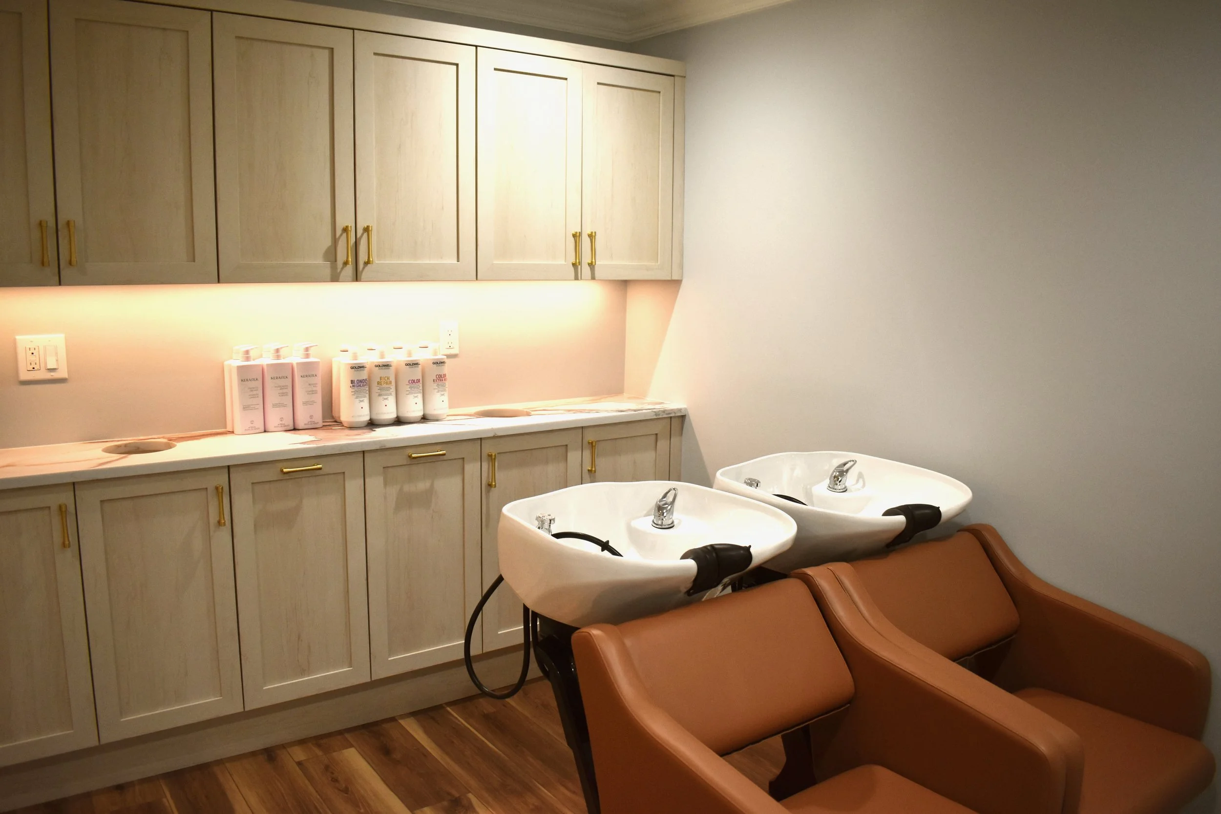 A salon or barbershop wash station with two brown reclining chairs, two white sinks with faucets, and a beige cabinet with six doors and gold handles above the sinks. Several bottles of hair products are lined up on the countertop underneath the cabinet.
