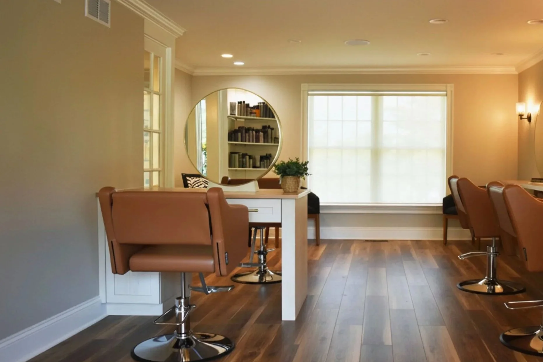A cozy room with a large window, a white desk with a plant, a circular mirror, bookshelves, and brown chairs on wooden floors.