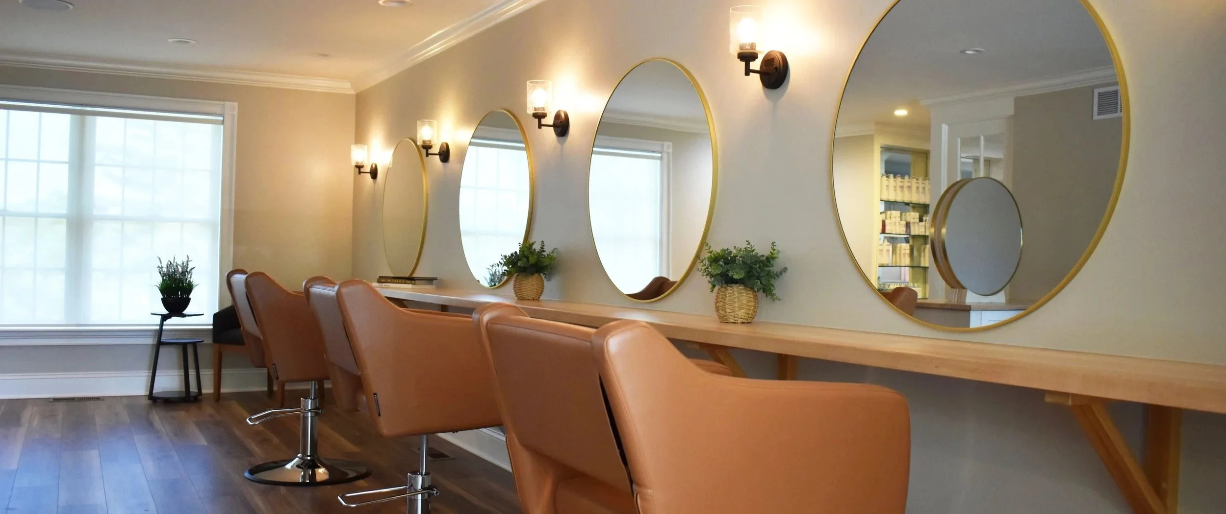 Salon or makeup station with large oval mirrors, tan chairs, small plants, and light fixtures on a beige wall.