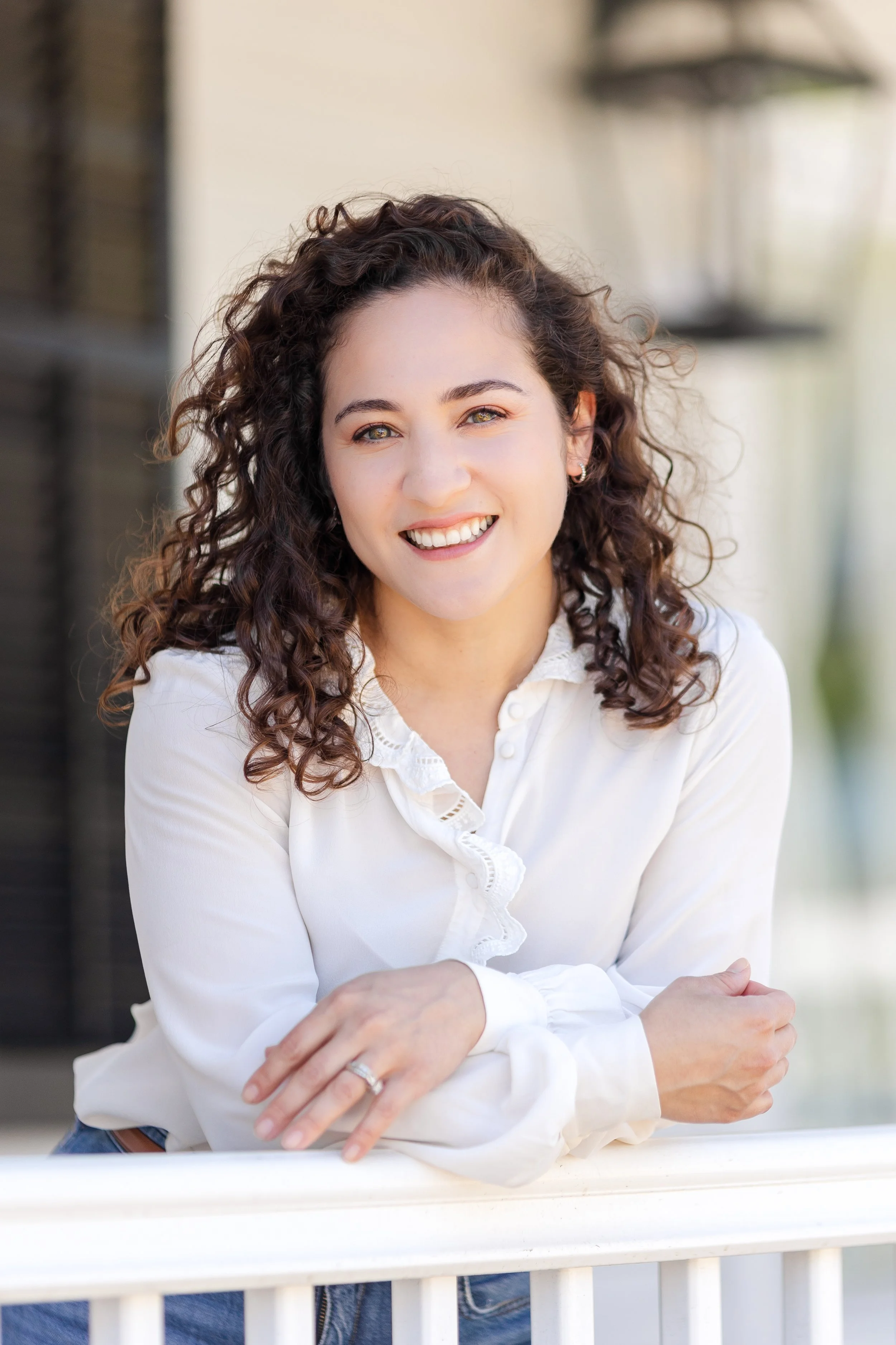 A woman with curly hair smiling and leaning on a white railing outdoors, wearing a white blouse and rings.