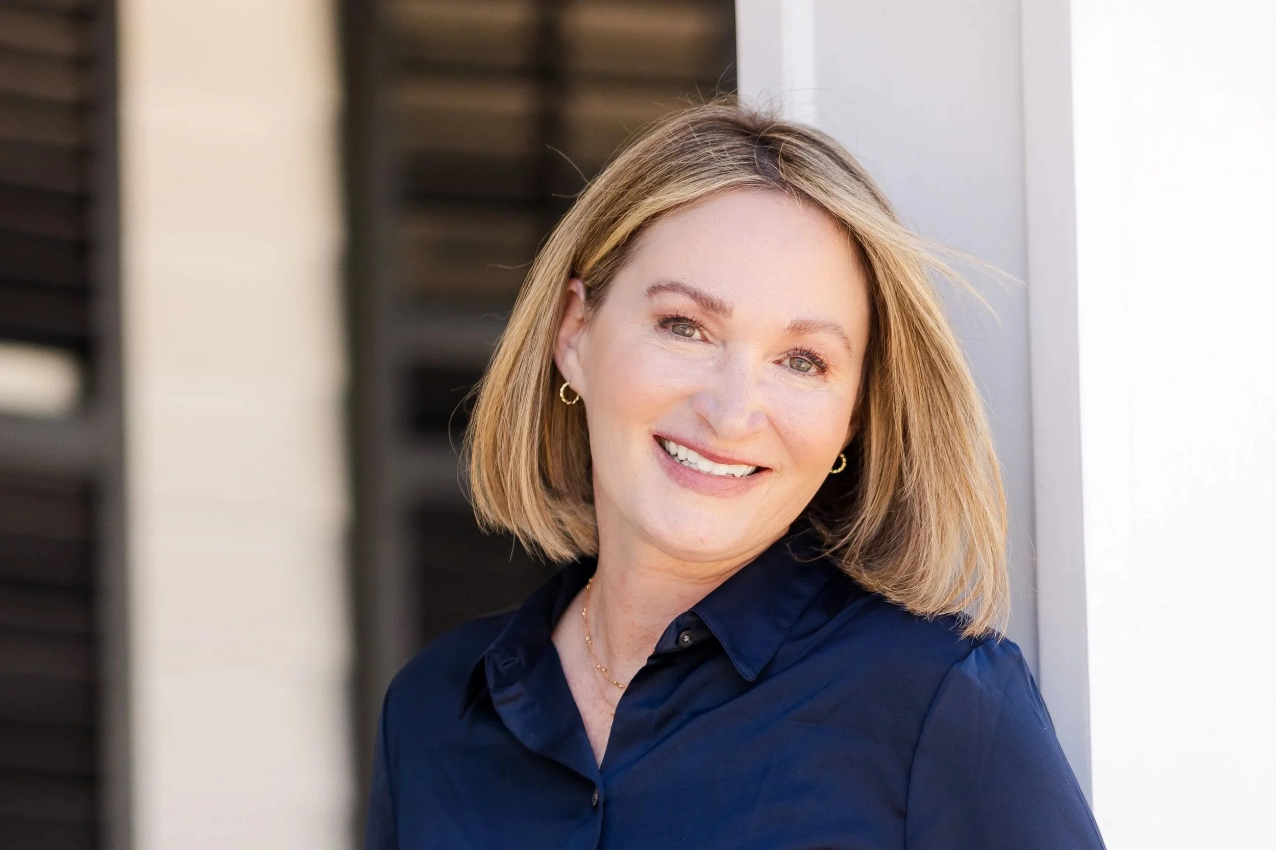 A woman with shoulder-length blonde hair, wearing a navy blue shirt with a collar, smiling and looking slightly to the right of the camera, standing outdoors near a building with white and dark window shutters.
