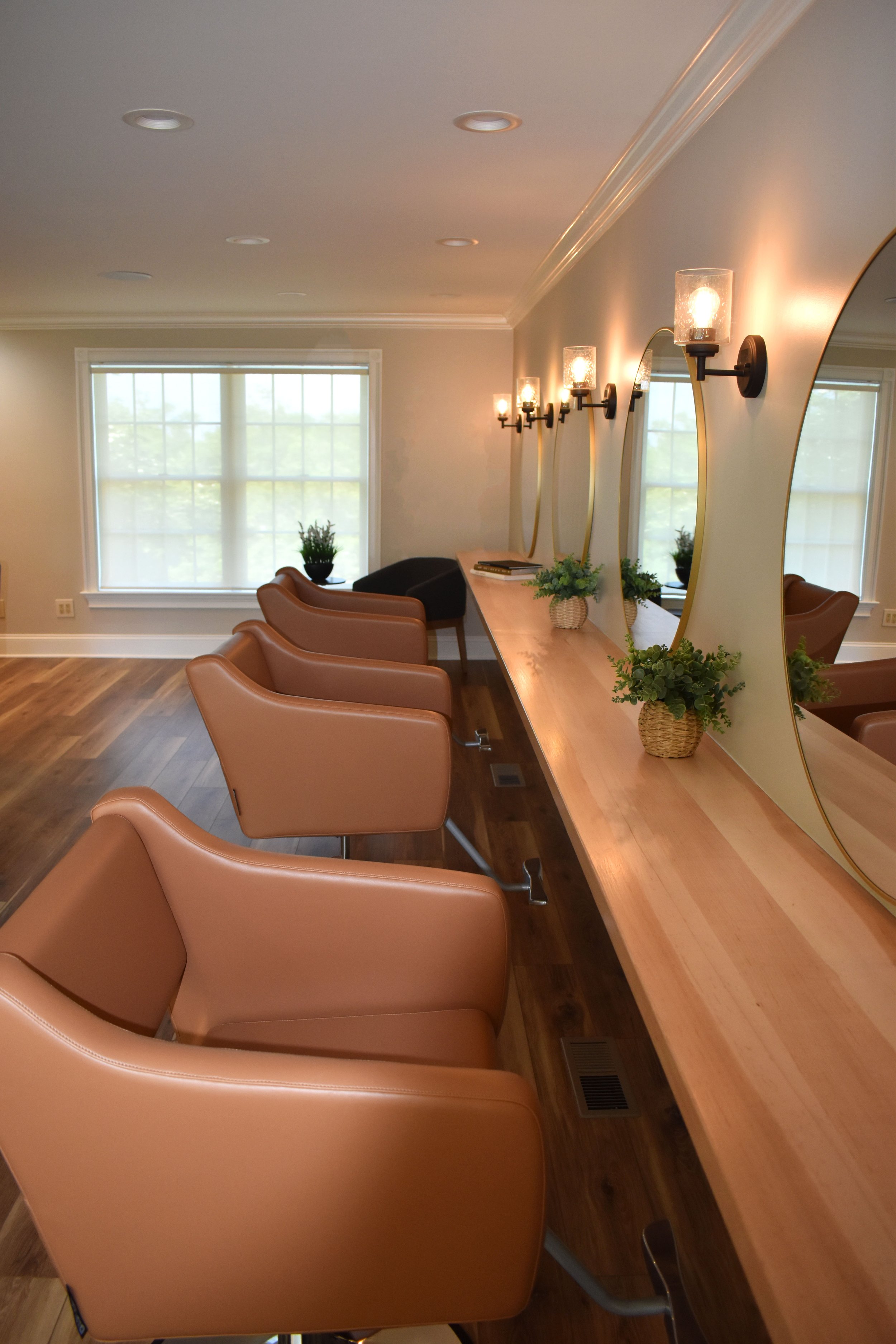 Interior of a room with brown seating, large windows with white curtains, a wooden table with potted plants and books, and framed mirrors on the wall with wall sconces.