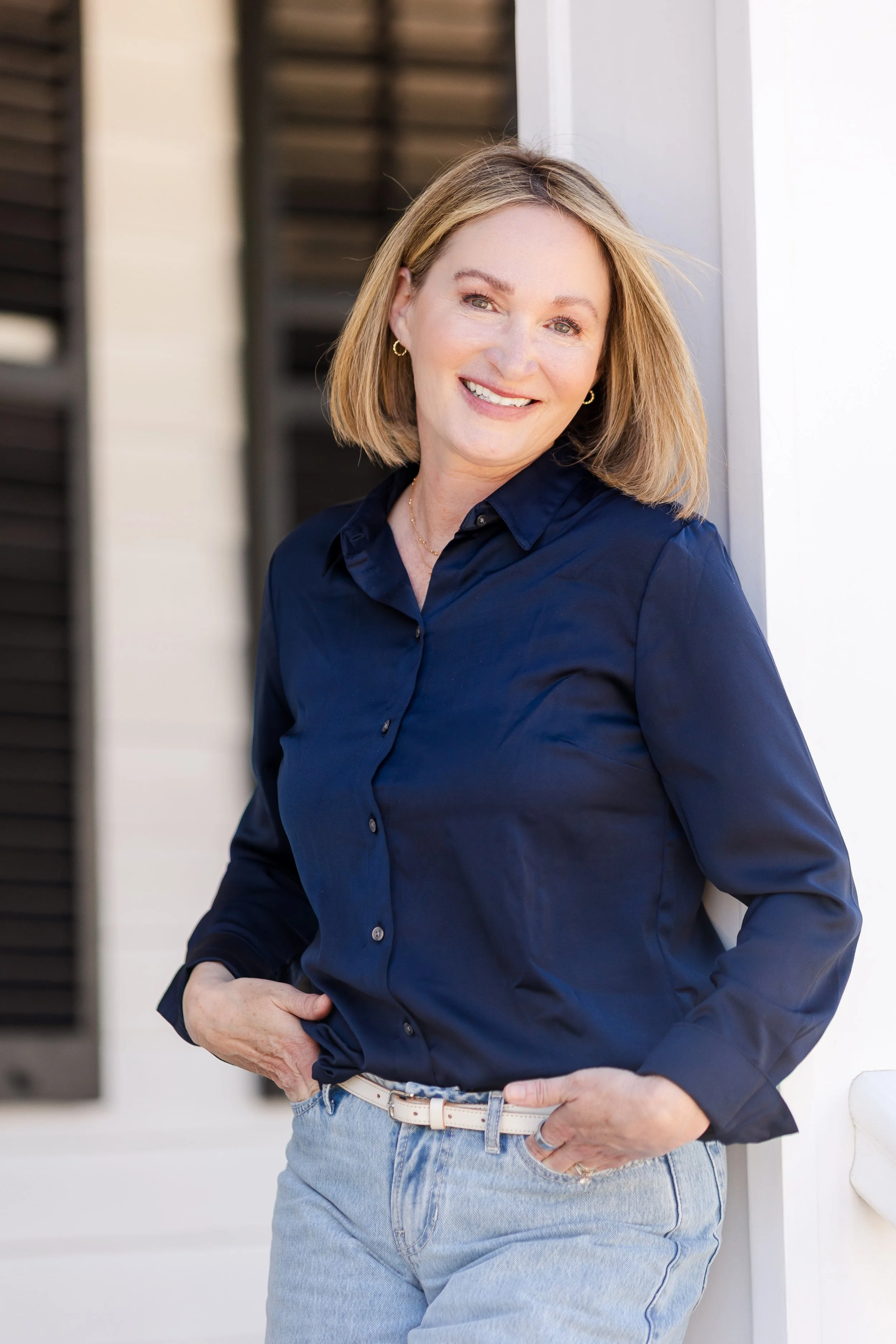A woman with shoulder-length blonde hair, wearing a navy blue button-up shirt and light blue jeans, leaning against a white wall outside in sunlight, smiling at the camera.