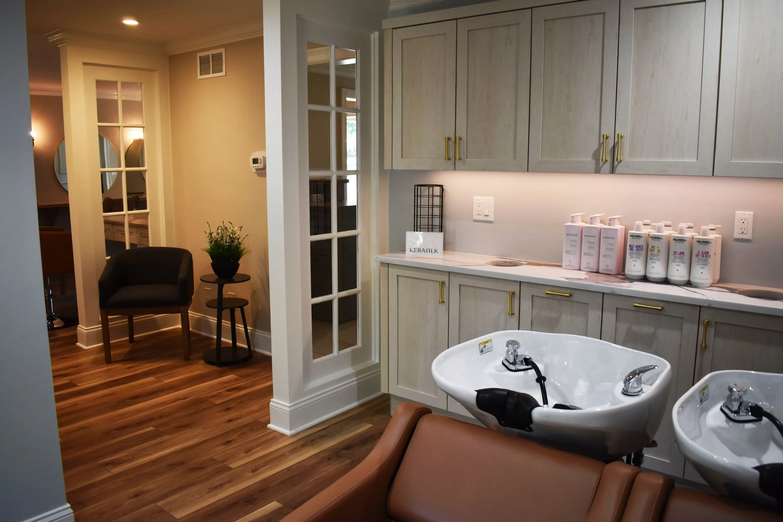 A salon or spa room with a wooden floor, beige walls, and a white cabinet with gold handles. There are two salon sinks with chairs, and a small table with a plant. In the background, there is a seating area with chairs and a patterned wall.
