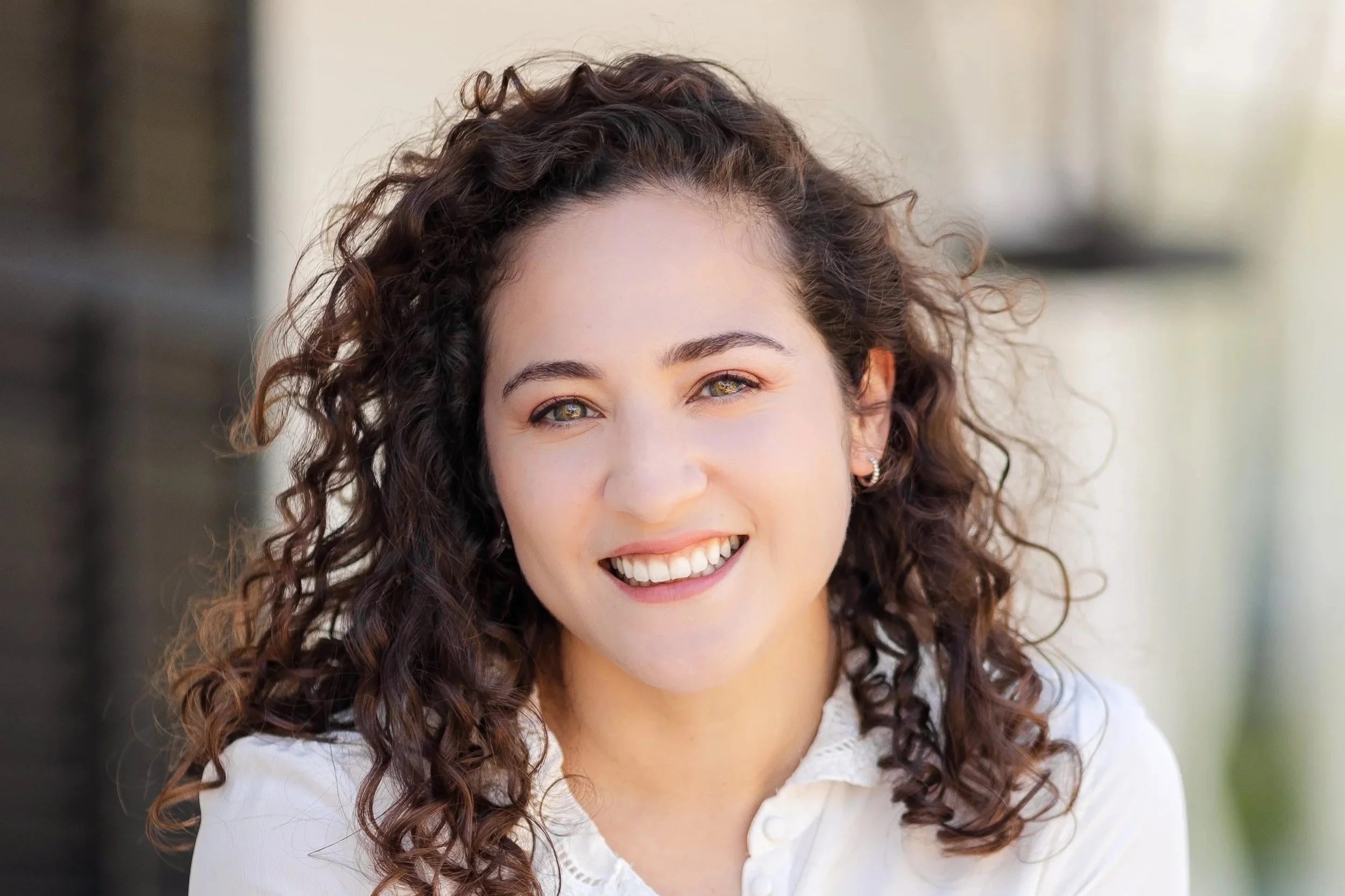 Close-up of a woman with curly brown hair, smiling, wearing a white top, with a blurred background.
