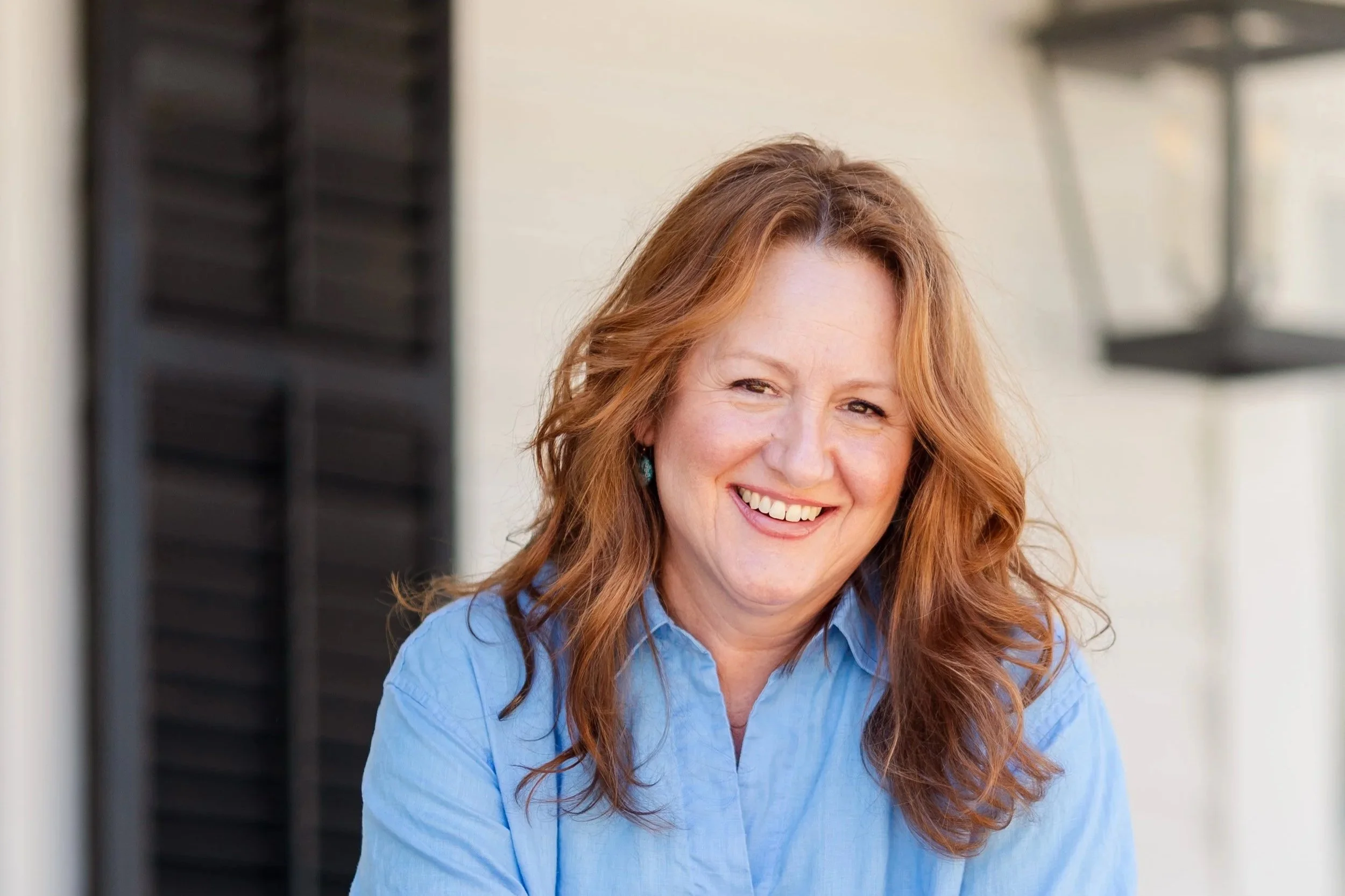 A smiling woman with long, wavy red hair wearing a blue shirt, standing outdoors near a white wall with dark shutters and a window in the background.