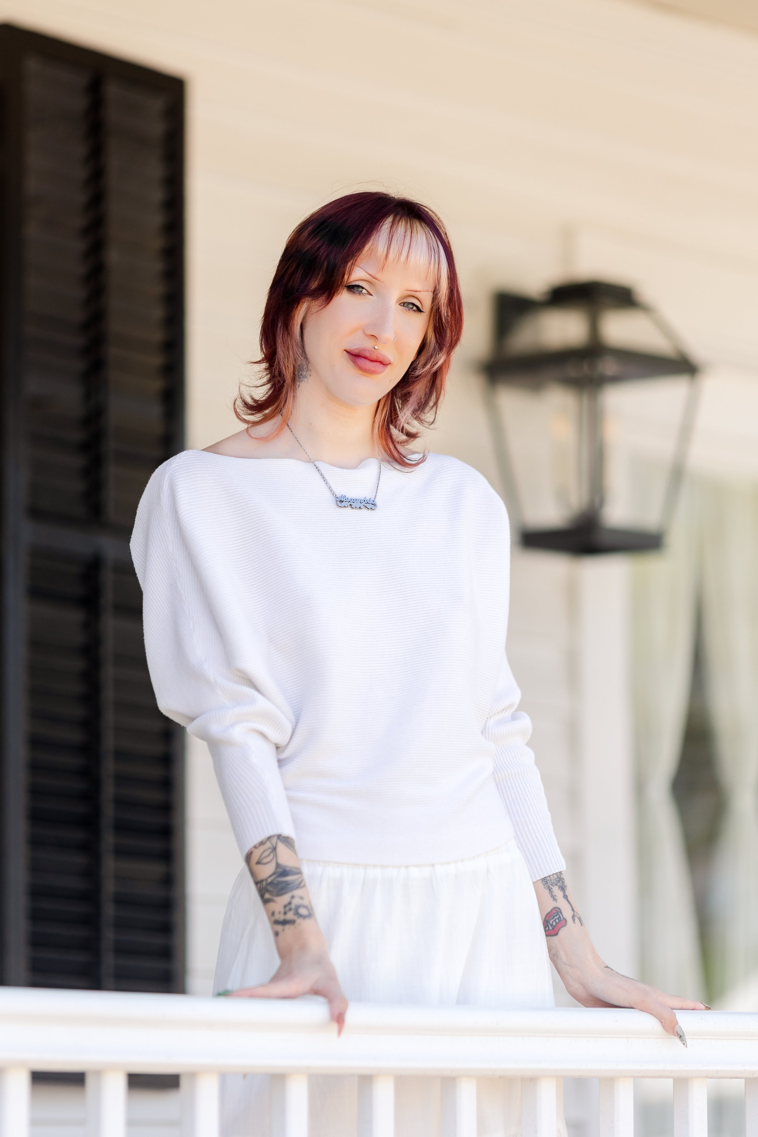 A woman with red hair and tattoos on her arms, wearing a white sweater and necklace, poses on a porch with a black wall and a lantern in the background.