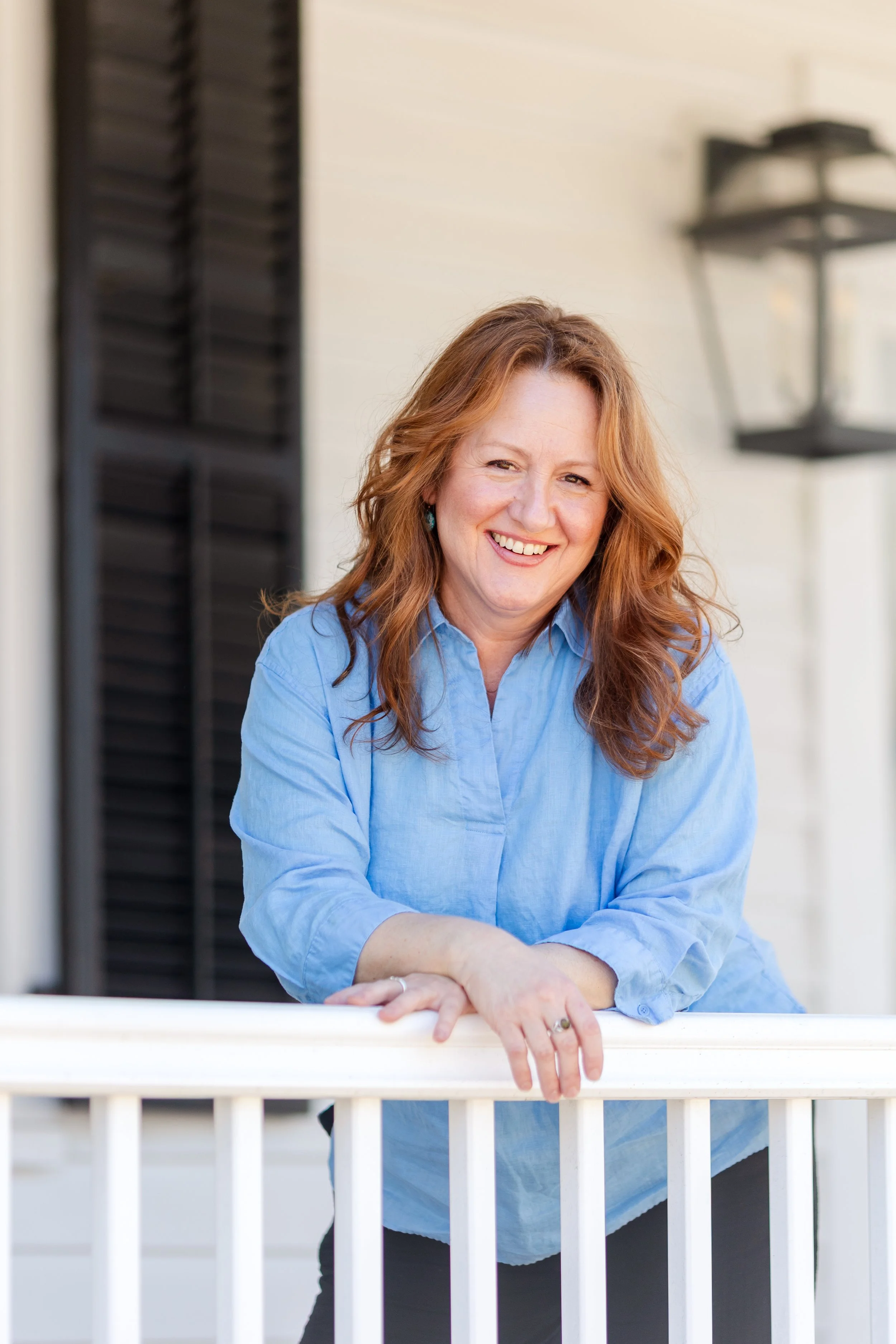 A woman with long red hair, wearing a blue button-up shirt, smiling and leaning on a railing on a porch or balcony, with a black shutter and a lantern in the background.