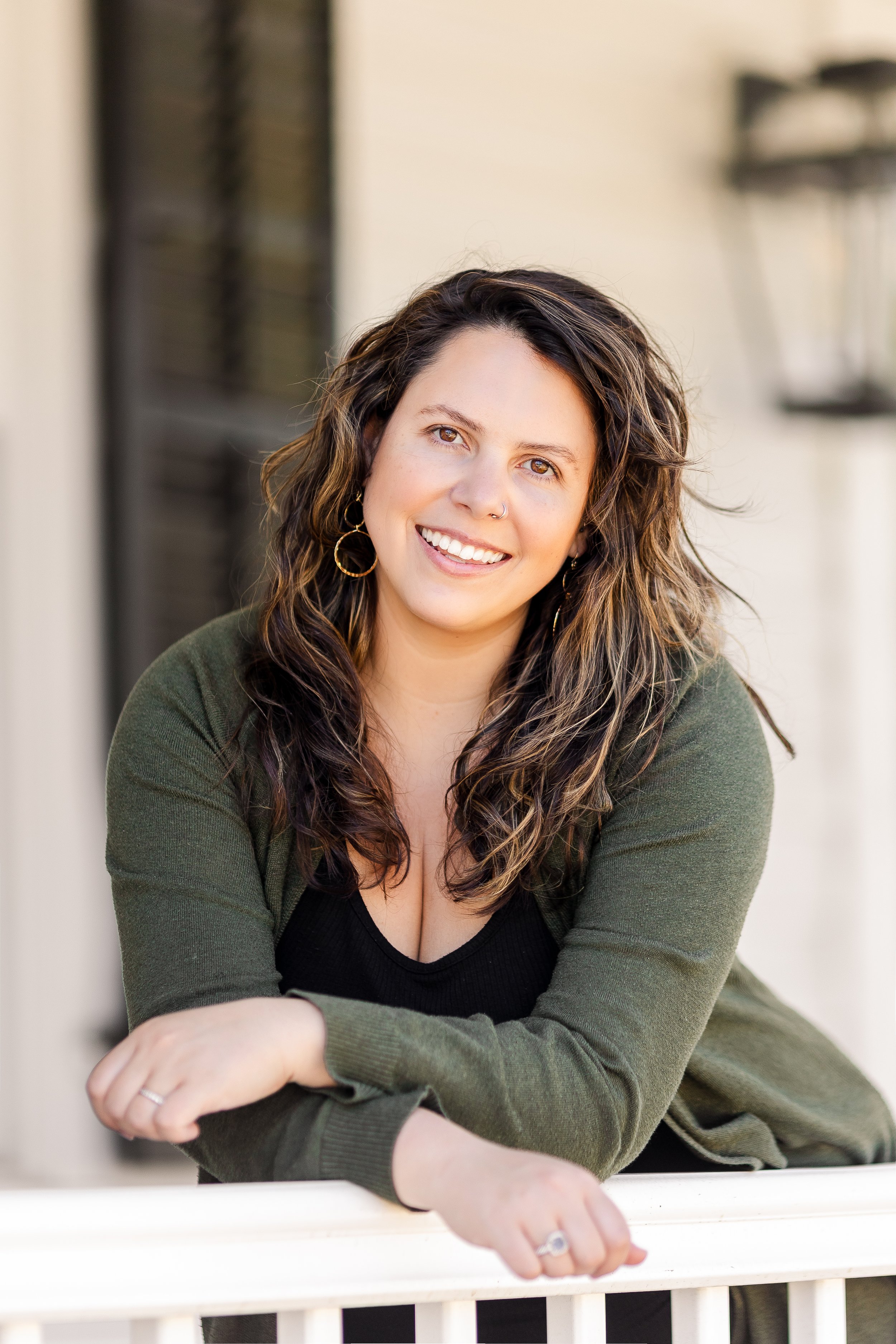 A woman with wavy brown hair and hoop earrings, smiling, leaning on a white railing outdoors, wearing a black top and green cardigan.
