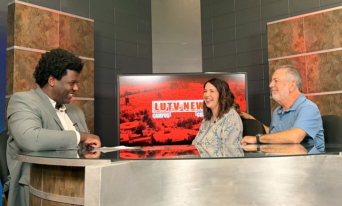 Three people sitting at a news desk, smiling and laughing during a TV show called 'LULU NEWS'.