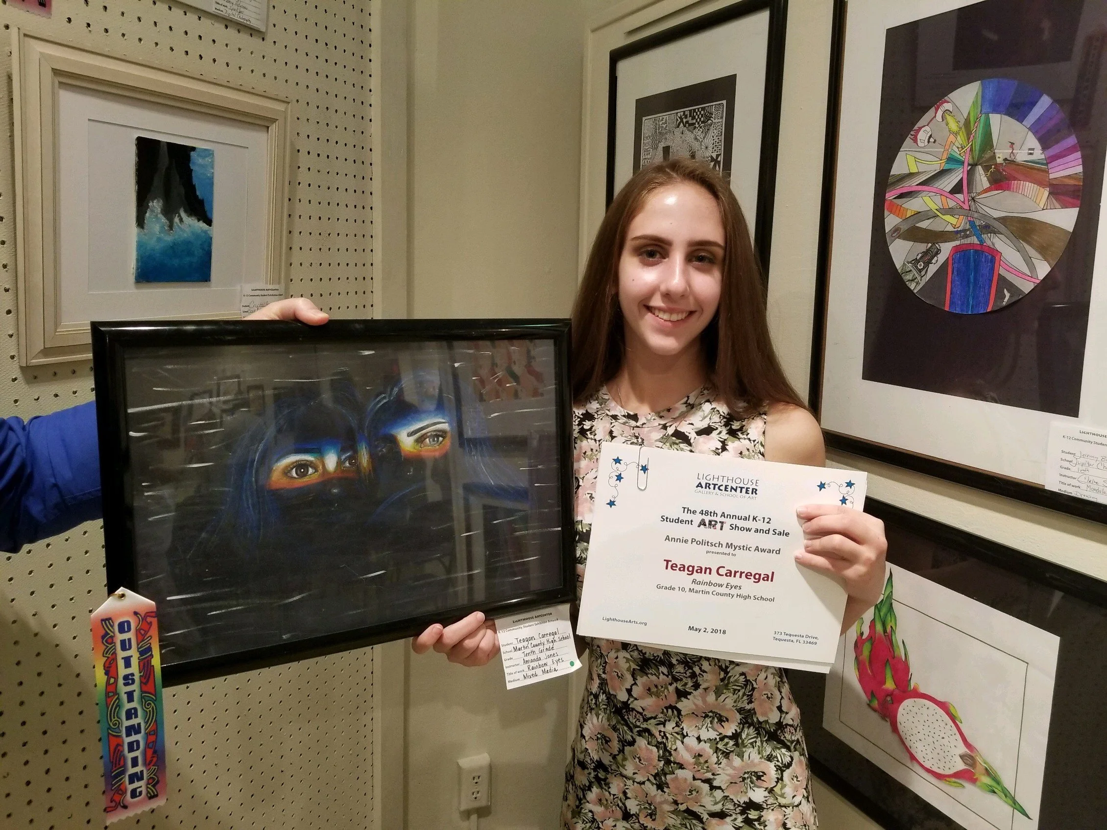 A young woman with long brown hair smiling, holding a certificate and a framed artwork, standing in an art gallery with various framed art pieces on the wall behind her.