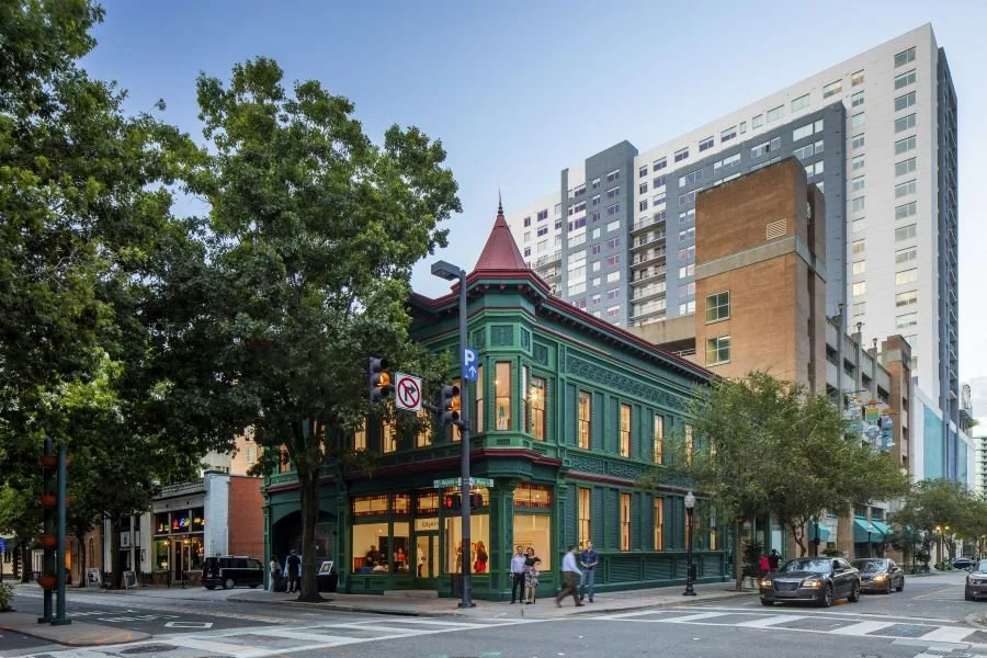 Historic green two-story building with large windows and a turret, situated at a street corner in an urban area with modern high-rise buildings and trees, during evening.