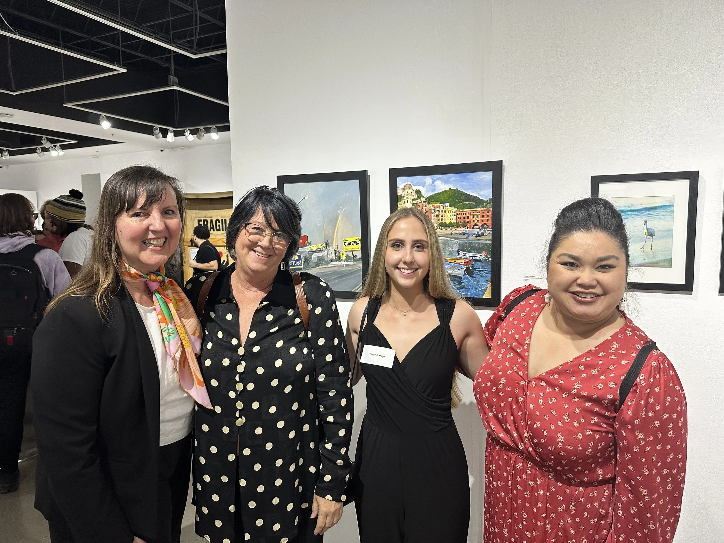 Four women standing side by side indoors at an art gallery, smiling at the camera, with framed paintings on the wall behind them.