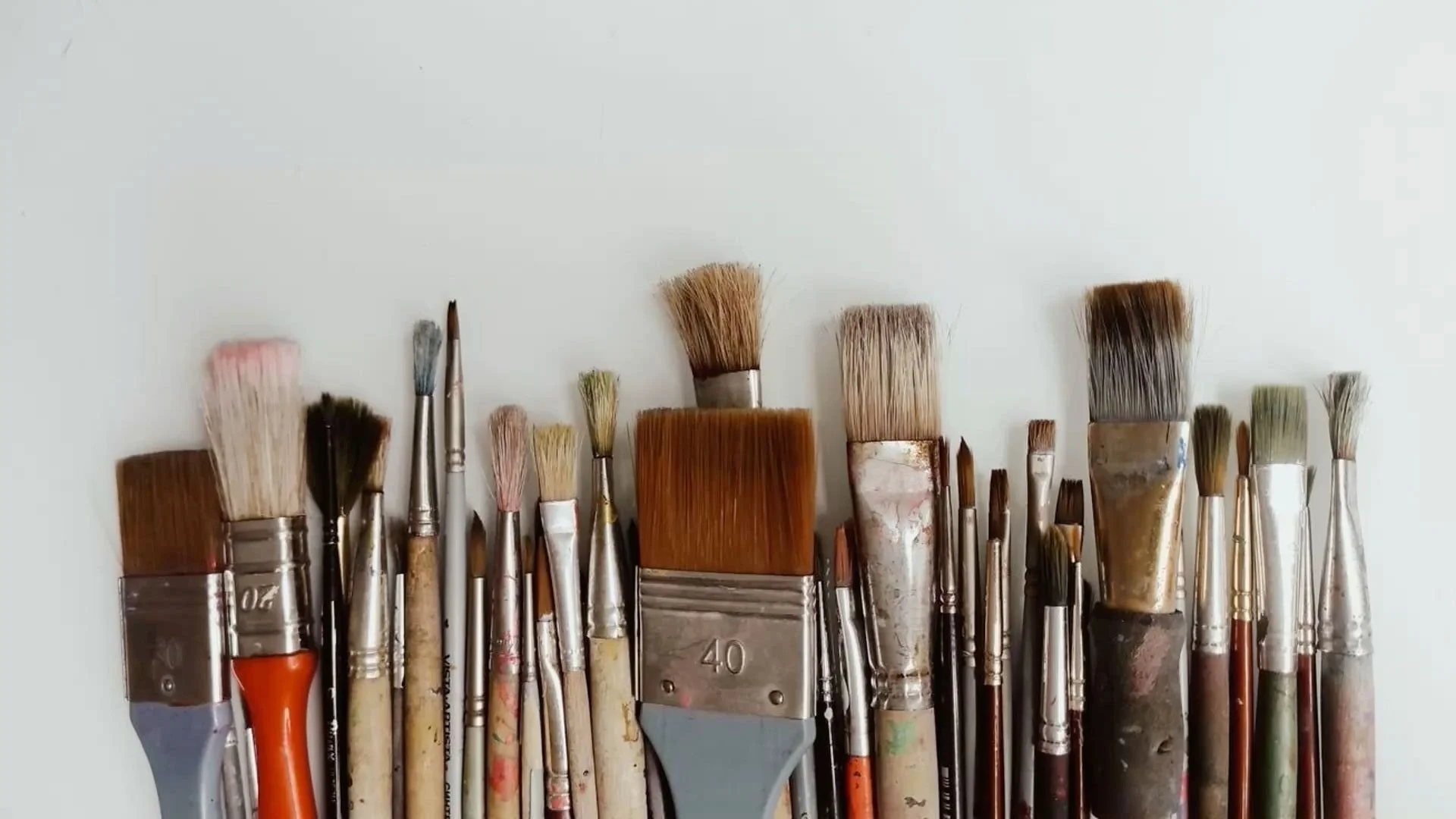 Various paintbrushes arranged horizontally, some with paint on bristles, of different sizes and shapes, with a plain white background.