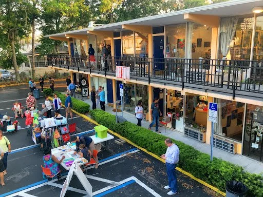 People gathered outside a two-story commercial building with a parking lot and outdoor tables.