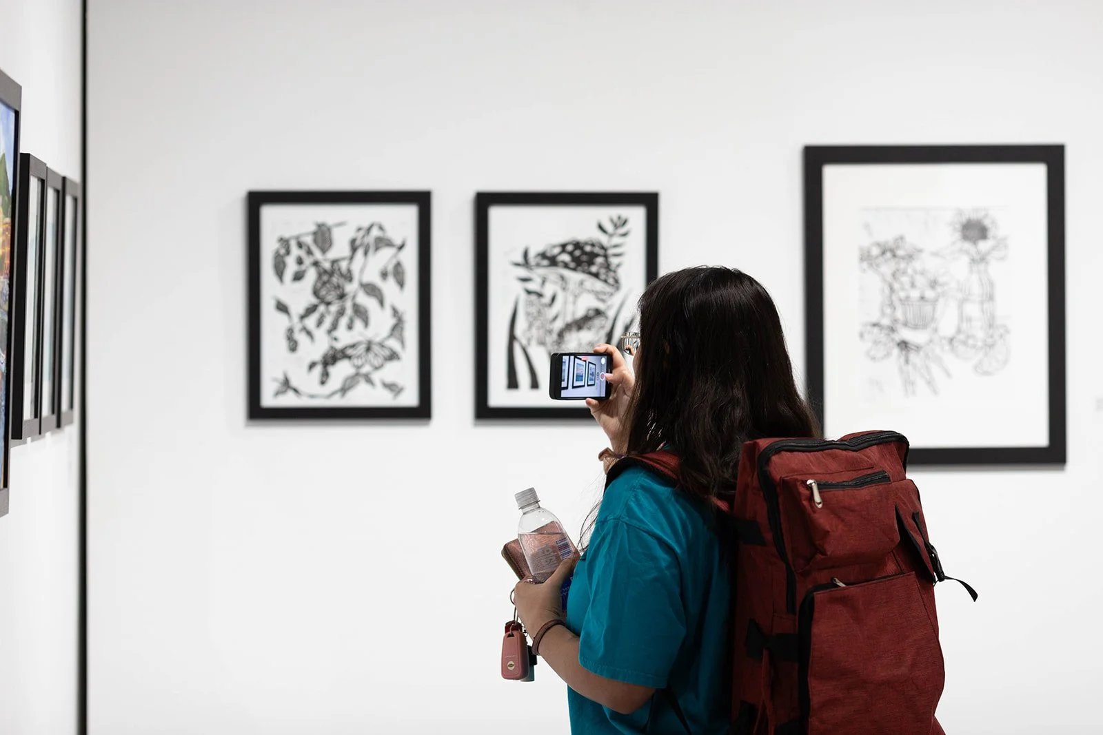 A woman with dark hair, wearing a teal shirt and carrying a red backpack, takes a photo of artwork in a gallery with her phone. She holds a water bottle in her left hand.