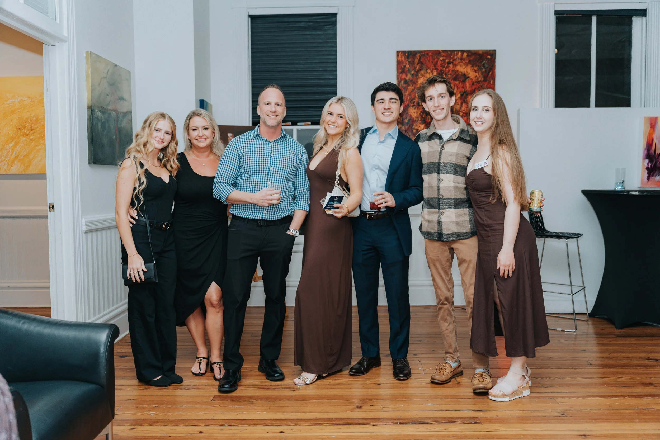 Group of seven young adults at an indoor social event, standing in a line and smiling at the camera, with artwork on the walls and a wooden floor.