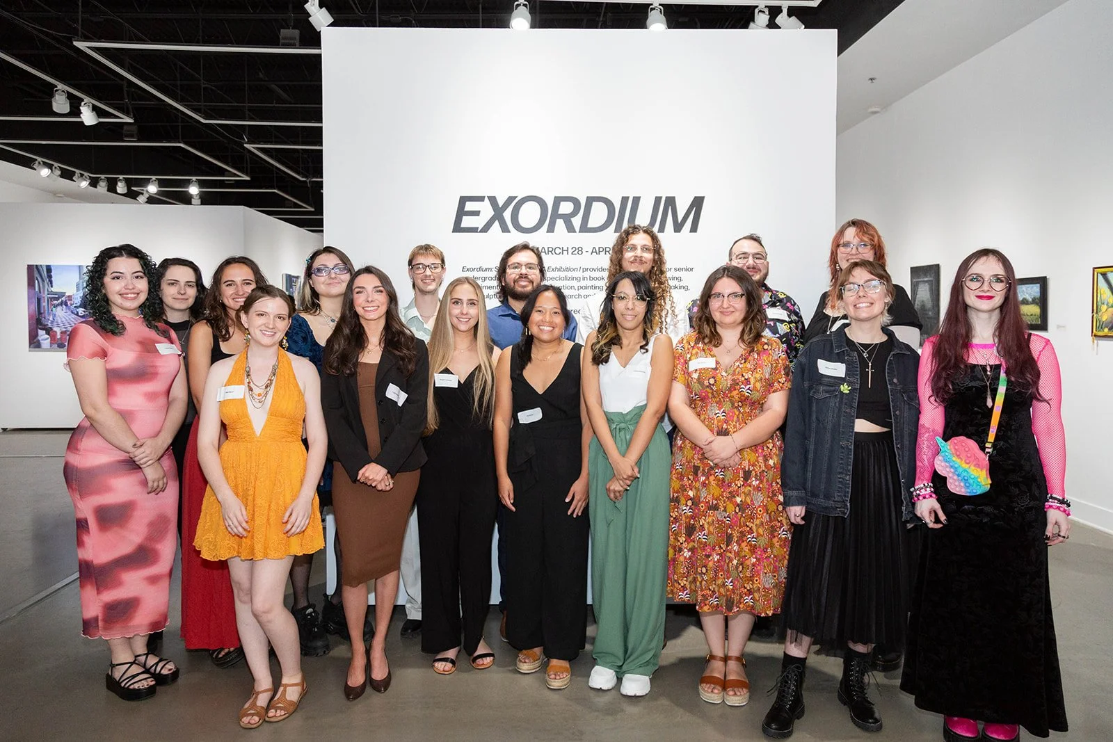 Group of people posing for a photo in an art gallery, standing in front of a white wall with a large sign that says 'EXORDIUM' and exhibition dates.