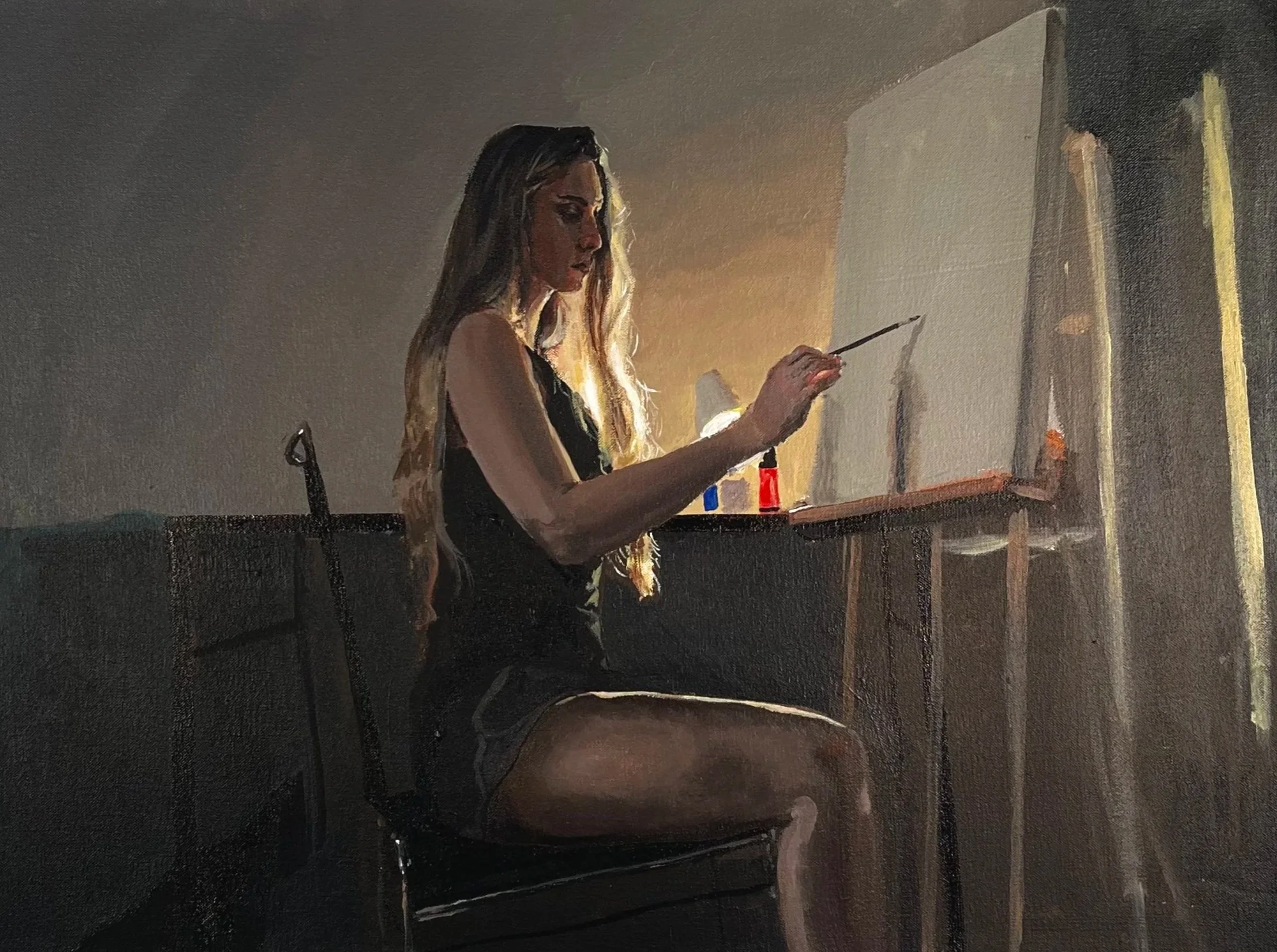 A young woman with long blonde hair painting on a canvas in a dimly lit room with a warm glow from a lamp, wearing a black sleeveless top and shorts, sitting on a chair.
