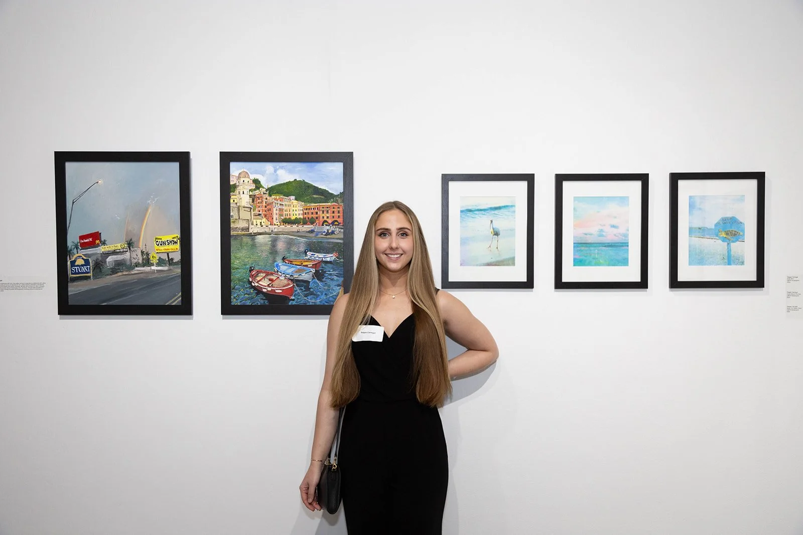 Young woman with long blonde hair, black dress, standing in front of framed paintings at the UCF art gallery.