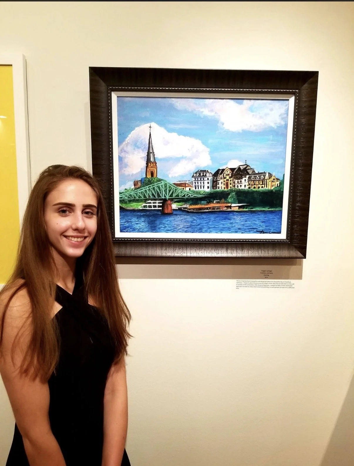 Young woman with long brown hair smiling in front of a framed painting of a cityscape by a river, featuring a prominent church spire, a green bridge, and colorful buildings against a blue sky with clouds.