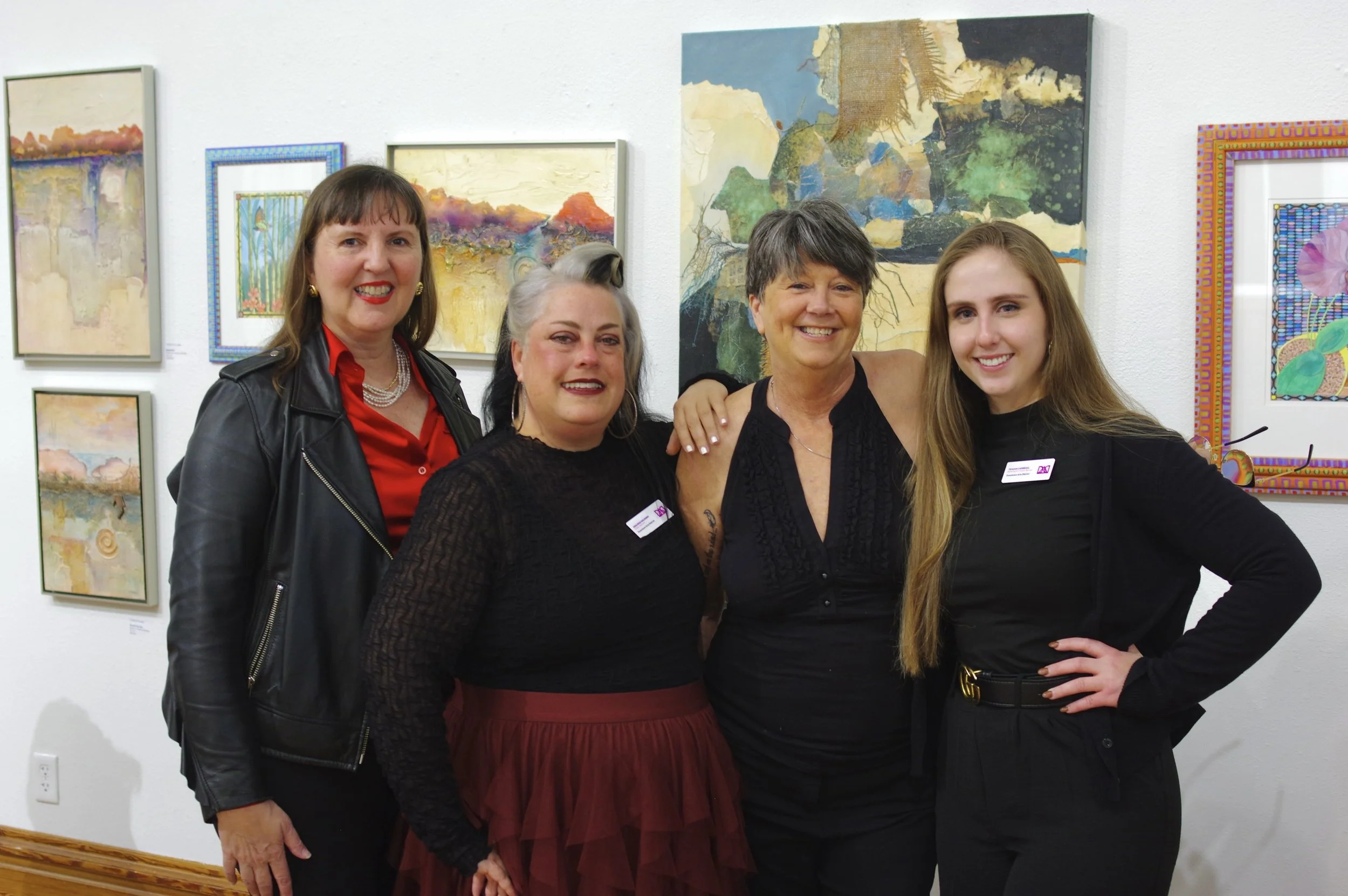 Four women smiling at an art gallery, standing close together, with colorful abstract paintings on the wall behind them.