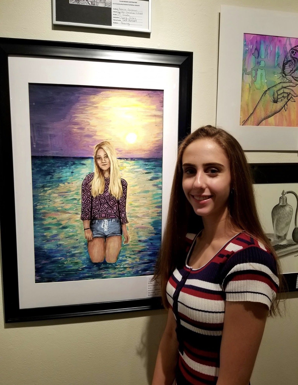 A young woman with long brown hair smiling at a colorful art exhibit next to a framed painting of a girl with blonde hair standing in water at sunset.