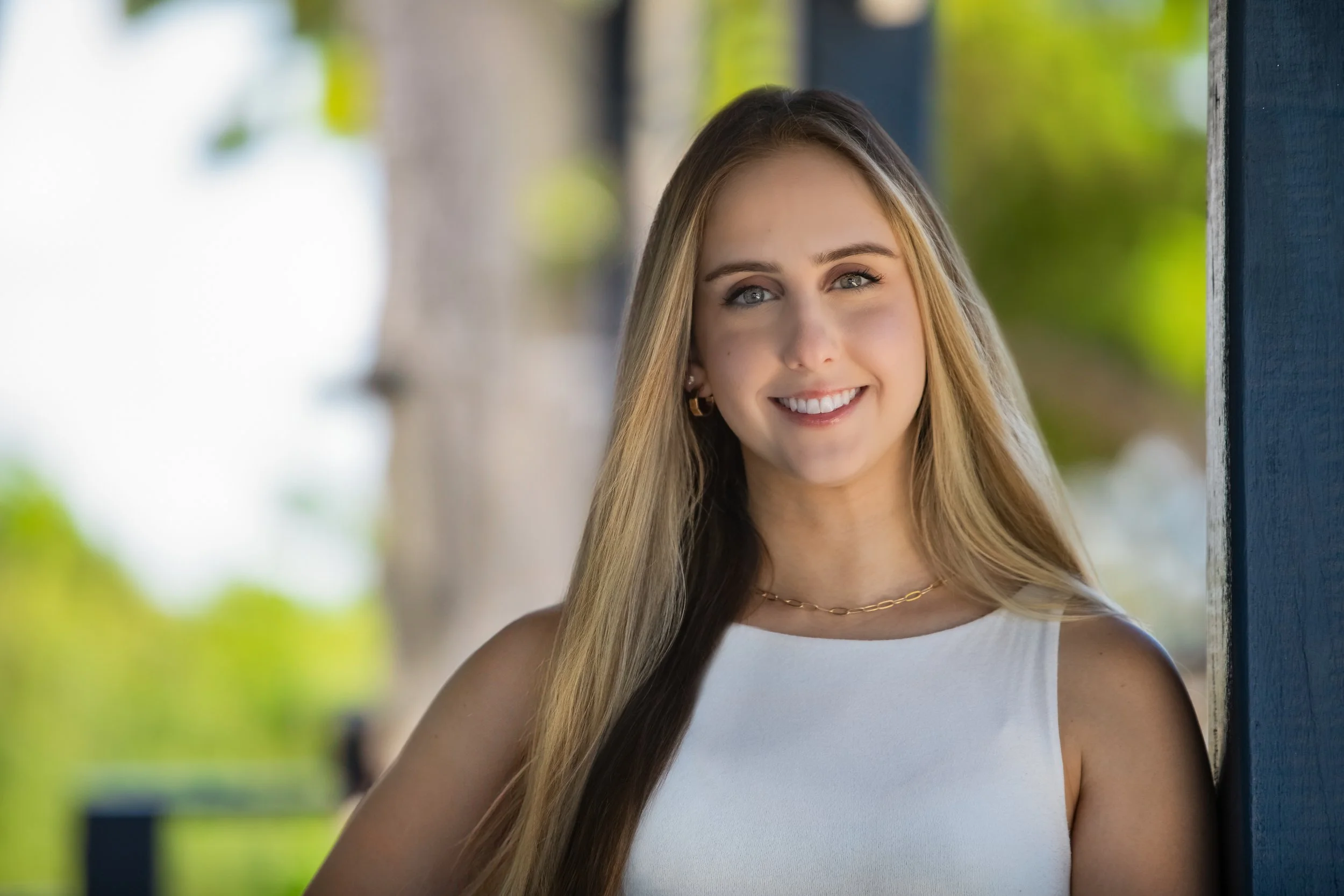 A young woman with long blonde hair and blue eyes smiling outdoors near a blue post, wearing a white sleeveless top and gold jewelry.