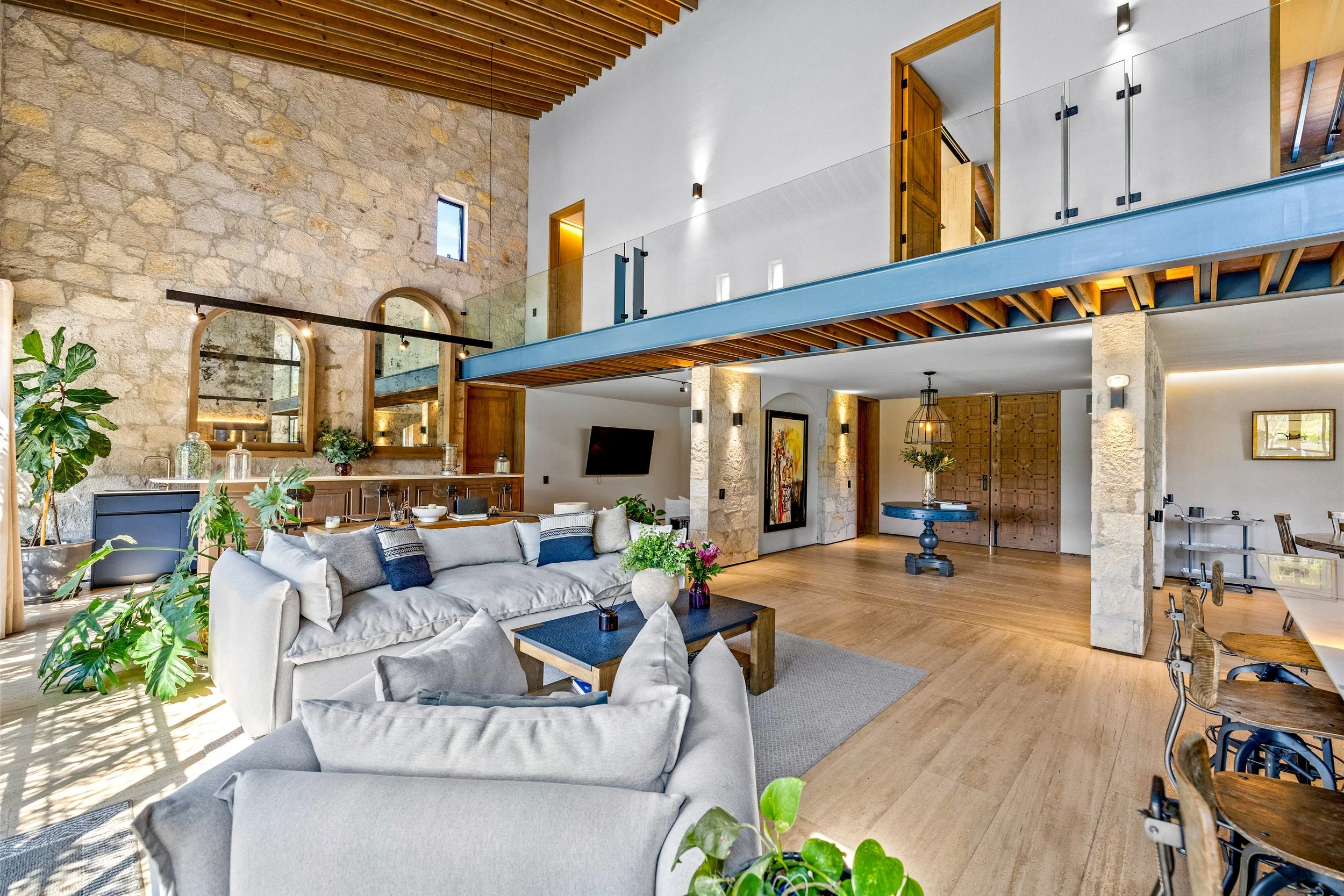 Spacious living room with stone walls, wooden ceiling, large windows, and a mix of modern and rustic furniture, including sofas, a coffee table, and decorative plants.