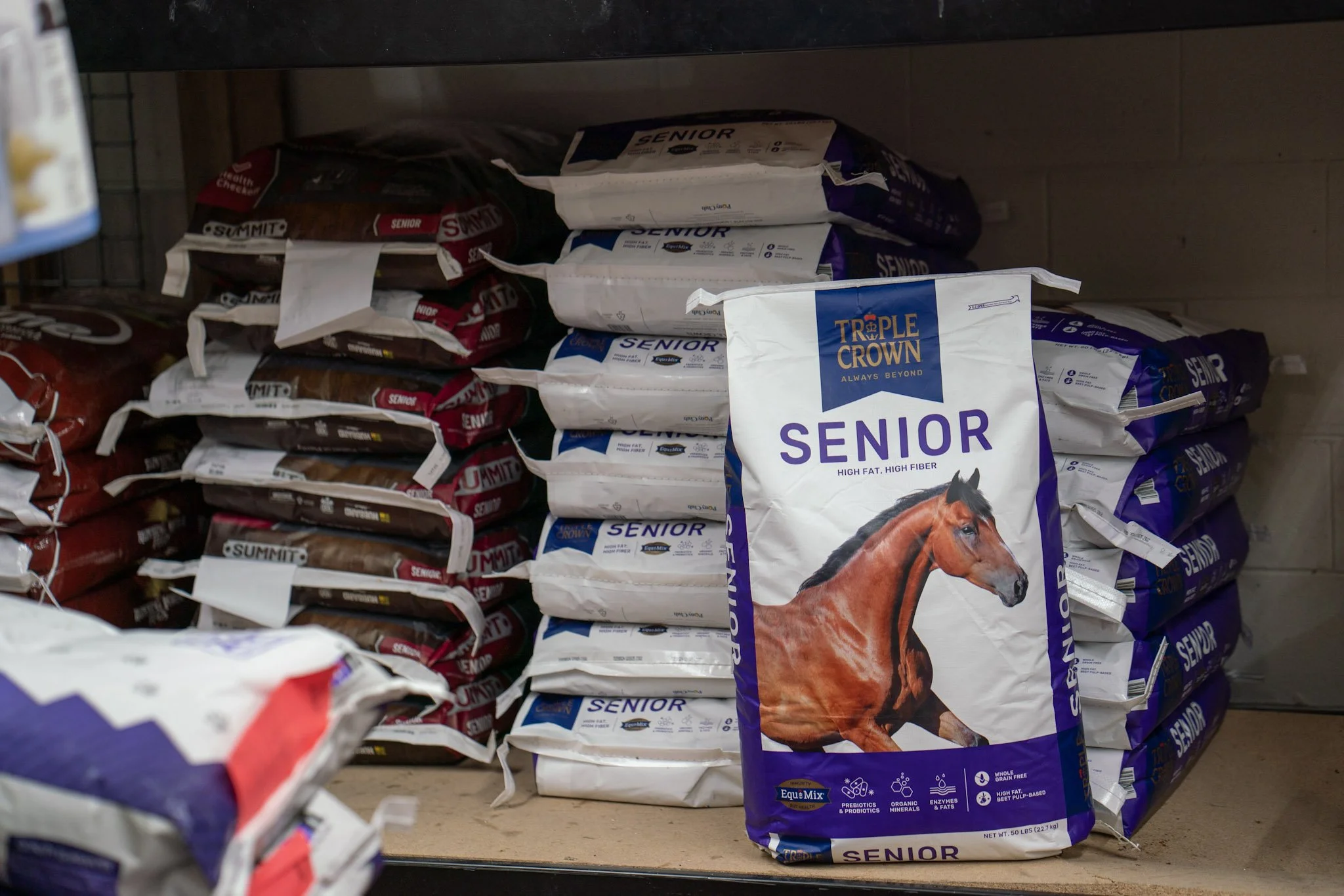 Shelf stocked with bags of horse feed, with a prominent bag labeled 'Senior' showing a horse image and brand 'Triple Crown' in a pet store or feed store.