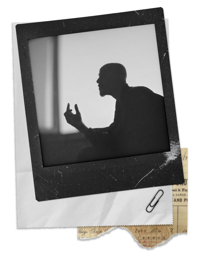 Silhouette of a man in profile, sitting and gesturing with his hand, in a black-and-white photograph.