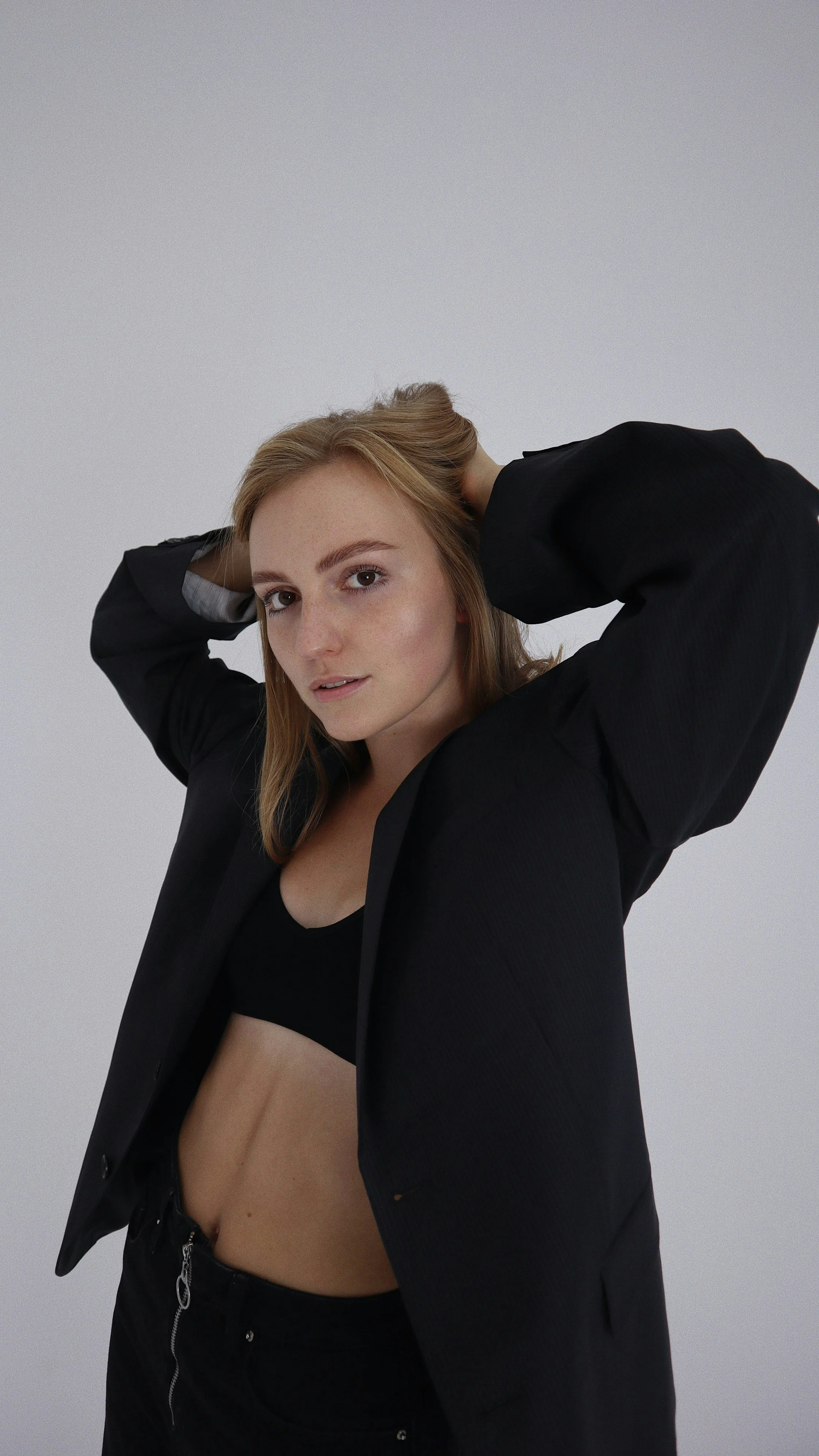 A woman with red hair wearing a black jacket, black crop top, and black pants, posing with both hands behind her head against a plain light grey background.