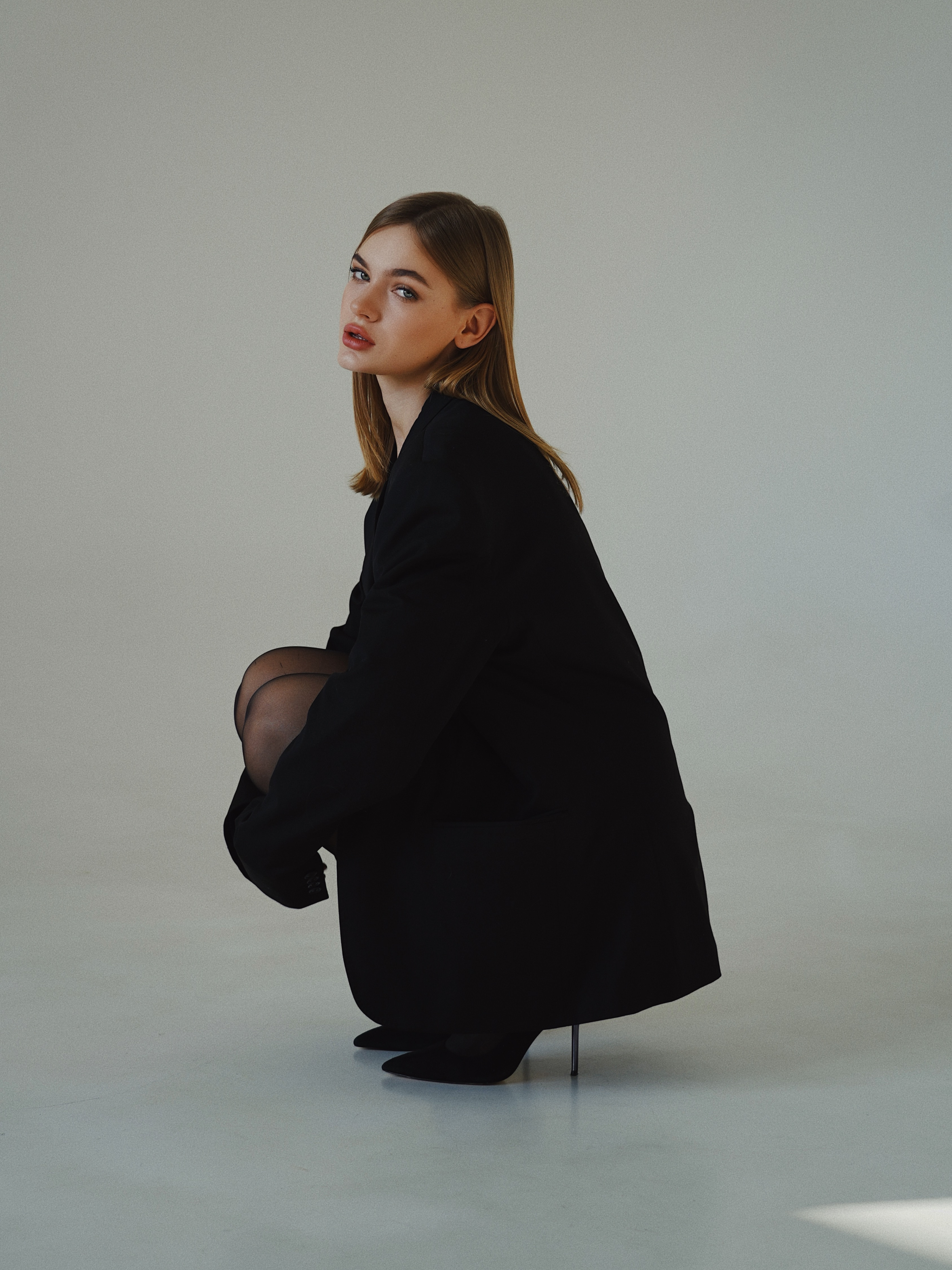 A woman with light brown hair and fair skin is squatting down, wearing a black oversized blazer and black pointed shoes, against a plain neutral background.