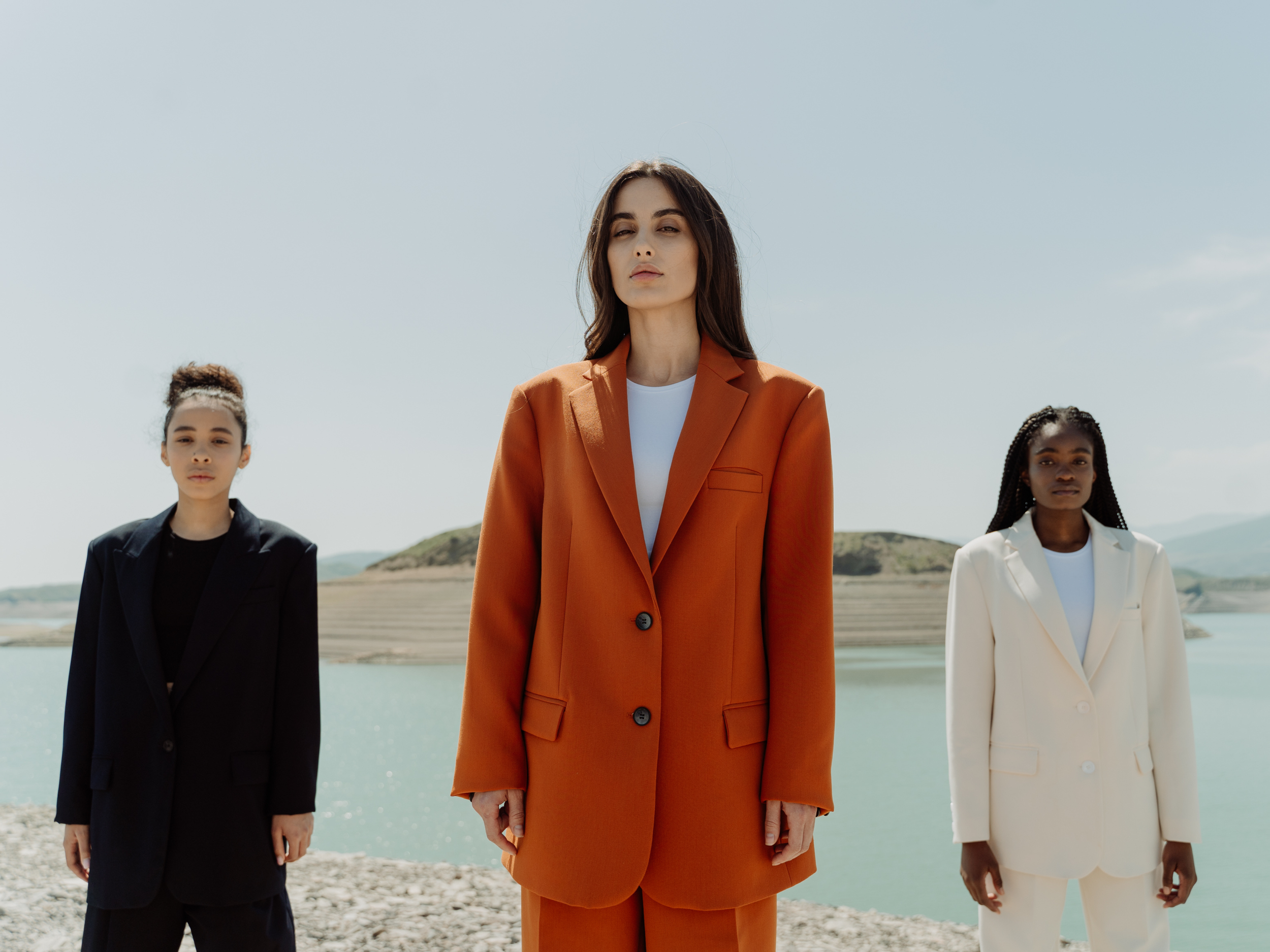 Three women standing outdoors by a body of water with hills in the background, wearing formal suits in black, orange, and white.