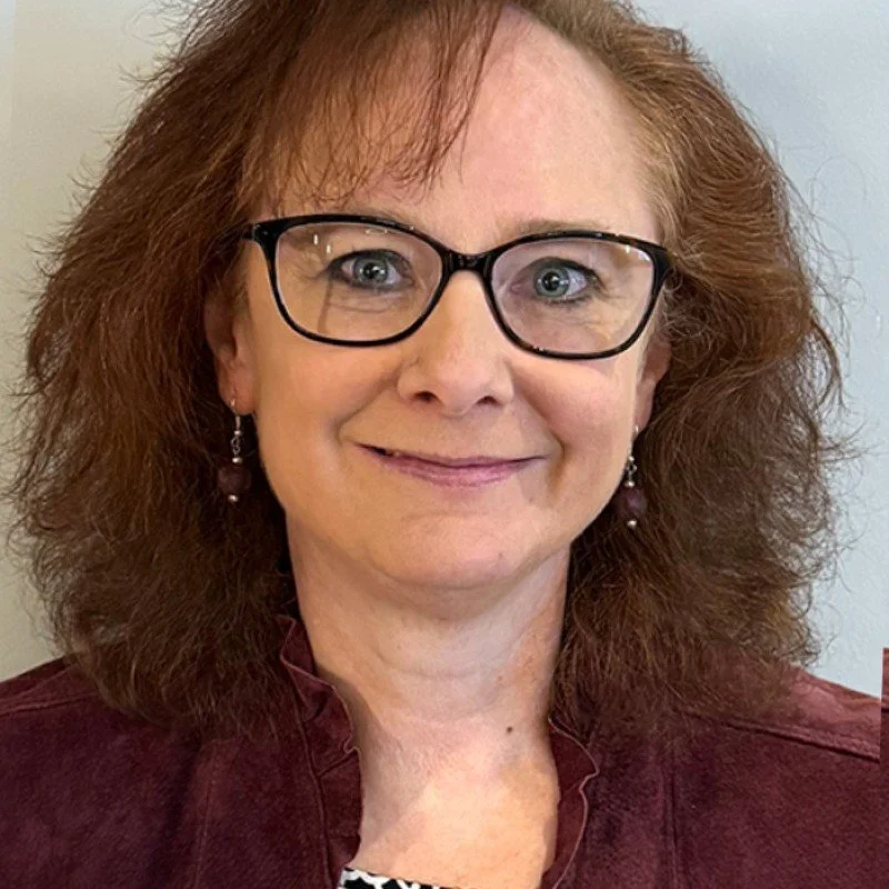 A middle-aged woman with red curly hair wearing glasses, earrings, and a burgundy jacket, smiling at the camera.