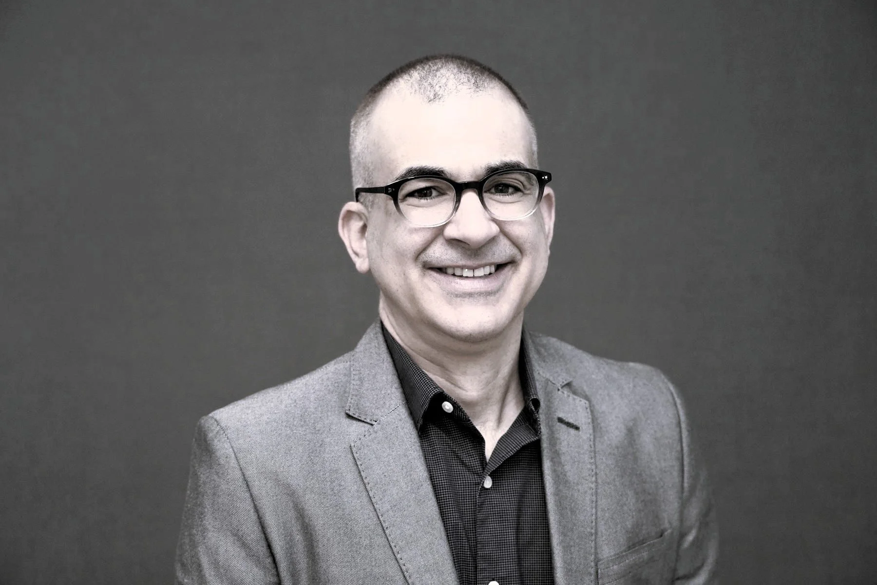 A smiling man with glasses, wearing a suit jacket over a button-up shirt, standing against a plain background.
