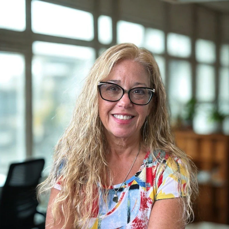 smiling woman with blonde curly hair, glasses, floral shirt, in a bright indoor space with large windows
