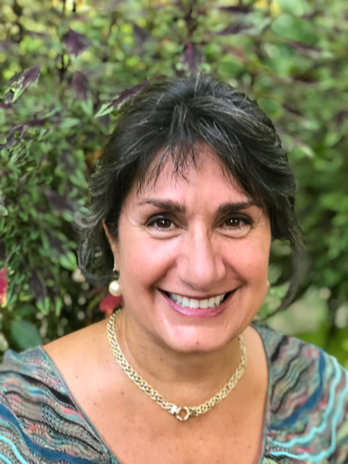 Smiling woman with short dark hair, wearing pearl earrings and a gold necklace, outdoors with green foliage in background.