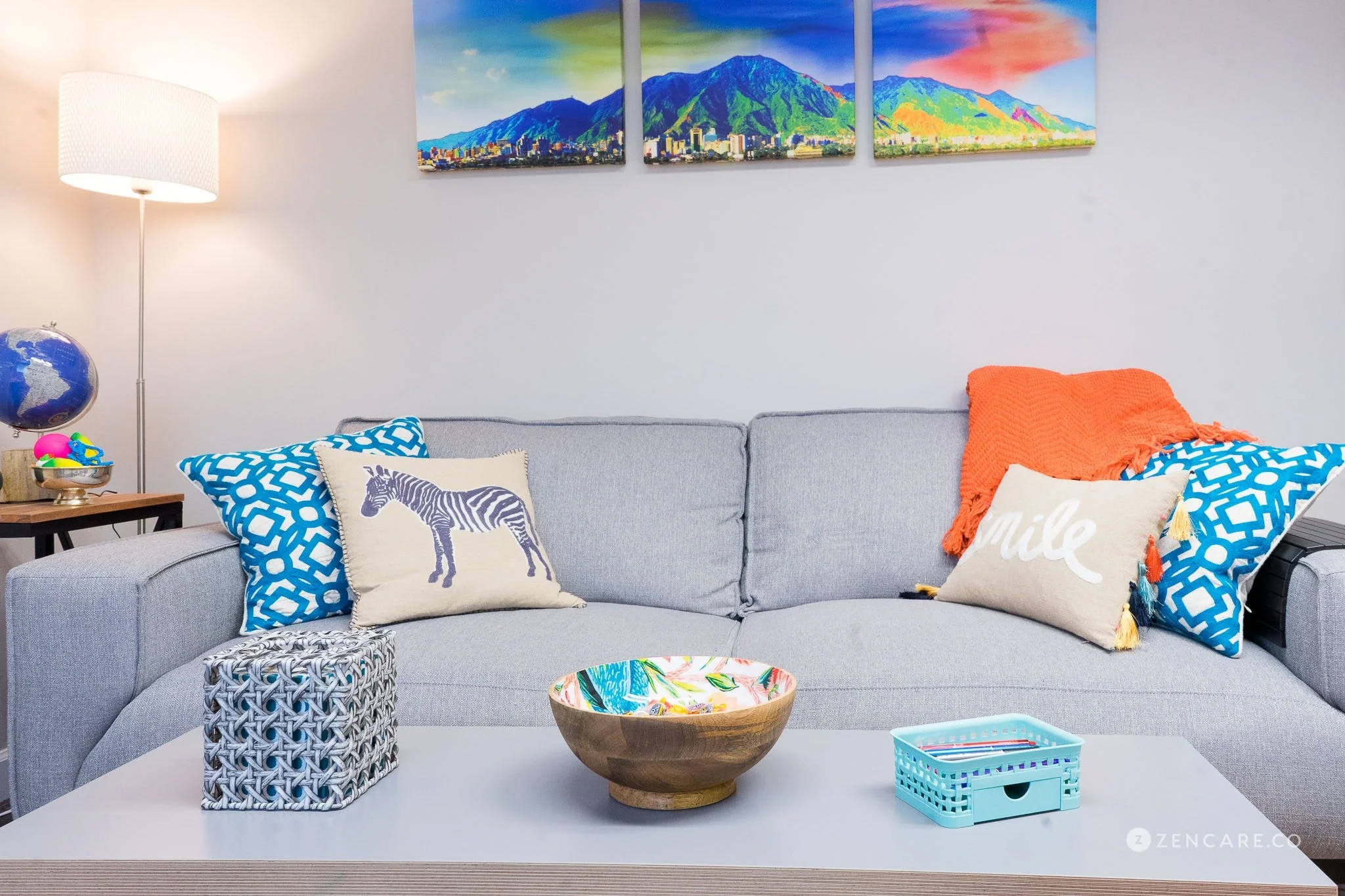 A living room features a light gray sofa decorated with colorful pillows, including one with a zebra print, another with the word 'Smile', and a blue geometric pattern. A bright orange throw blanket is draped over one side. The white coffee table in front holds a wooden bowl with a colorful interior, a woven tissue box, and a small blue storage basket. A tall floor lamp with a white shade stands next to the sofa. Behind the sofa, there is a wall art piece with a scenic mountain landscape divided into three panels.