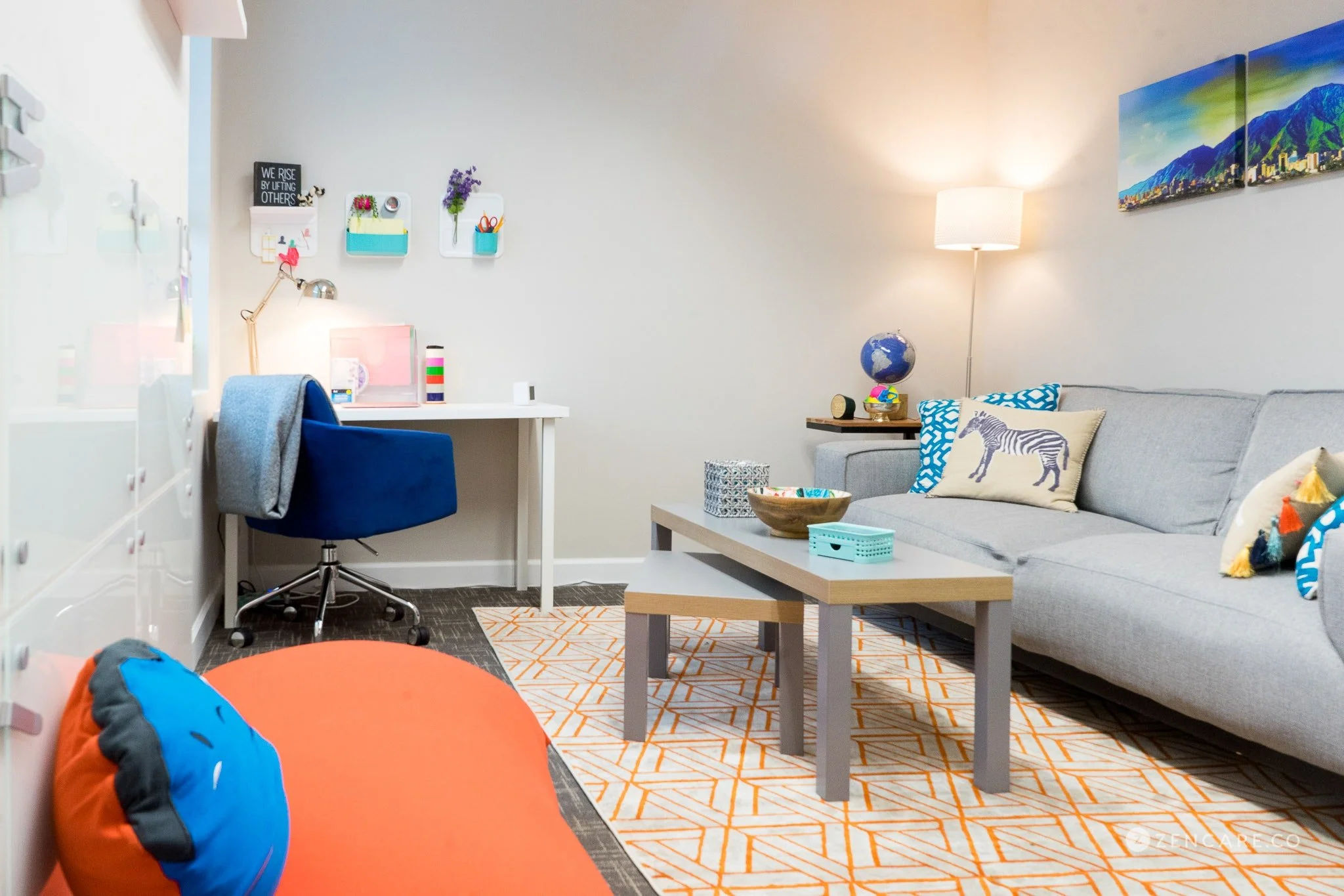 Living room with a gray sofa decorated with animal-themed pillows, a coffee table, and a table against the wall with a globe, stacked books, and a lamp. A wall art piece depicts a mountain scene, and a small desk with a blue chair is set up in the corner. The room has a patterned orange and white rug and various colorful decorations.