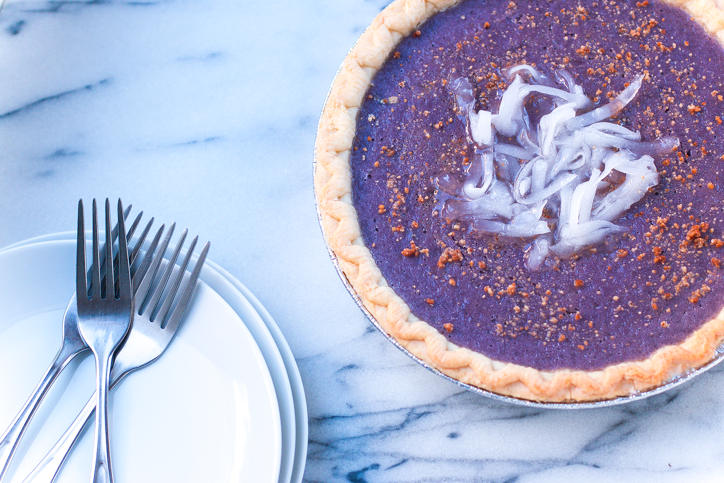 Ube pie with a golden crust, topped with Macapuno, on a marble surface, next to a stack of white plates and three silver forks.