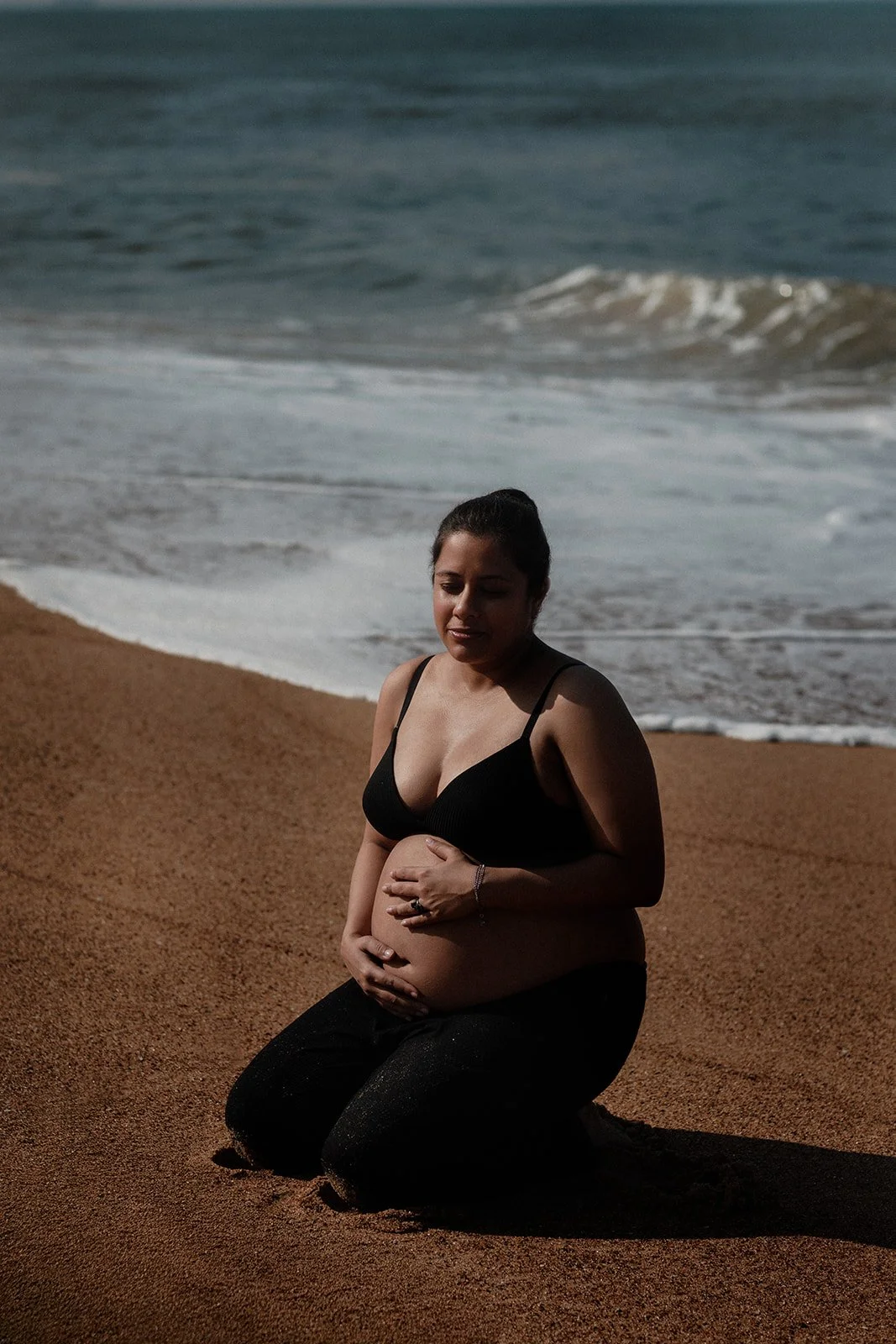 A pregnant lady kneeling on the beaches holding her belly as she is being photographer by Darius N Fernandes