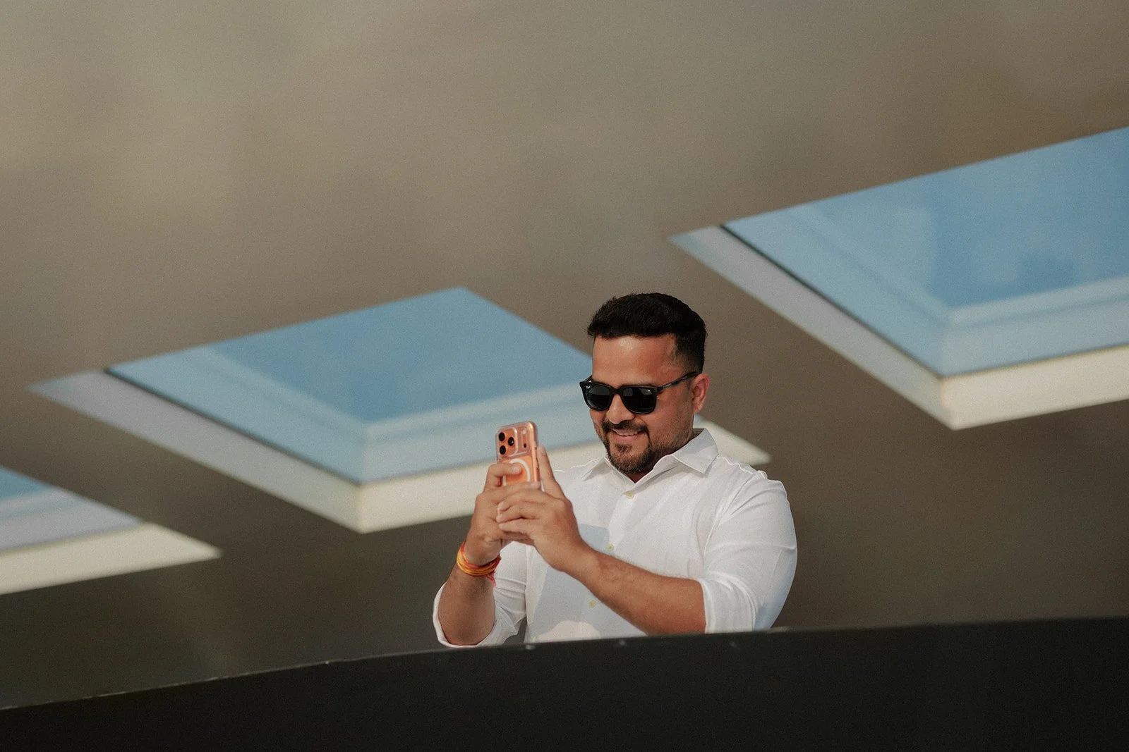 event photograph of a Man in white shirt and sunglasses smiling and looking at his phone as he takes a picture under skylights.