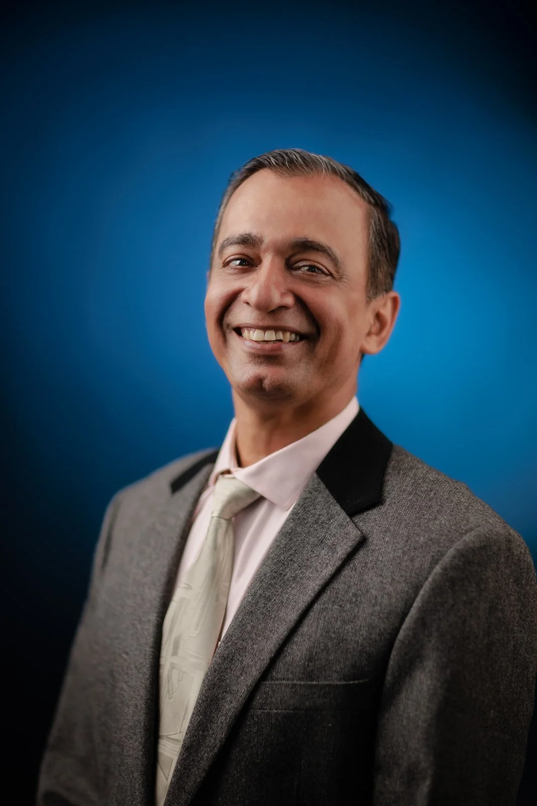 A corporate headshot photograph of a man in a grey suit with a blue background 