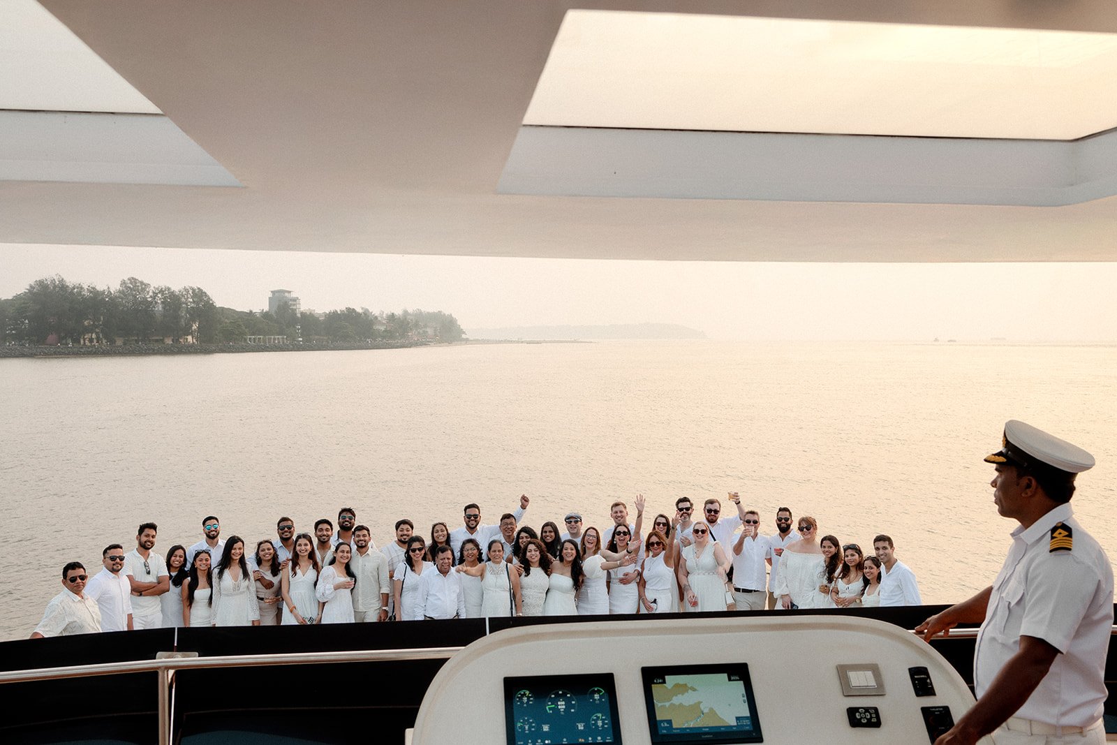 A group of people dressed in white for an event on a Yatch boat with a captain in uniform, near water with a distant shoreline and trees.