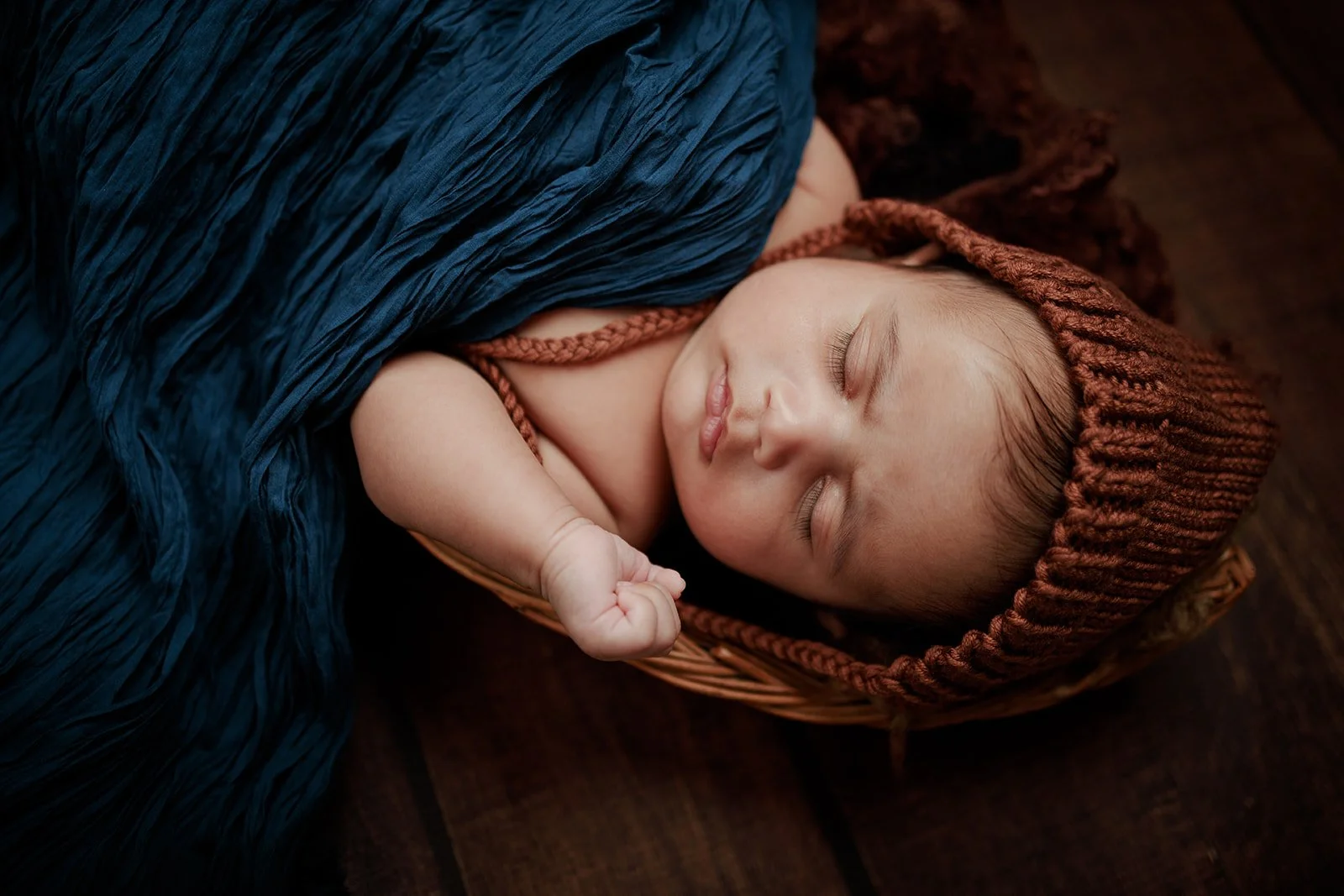 A photograph of a baby cuddled up on a wooden backdrop with a blue cloth draped on him and a brown beanie taken by new born photographer Darius N Fernandes