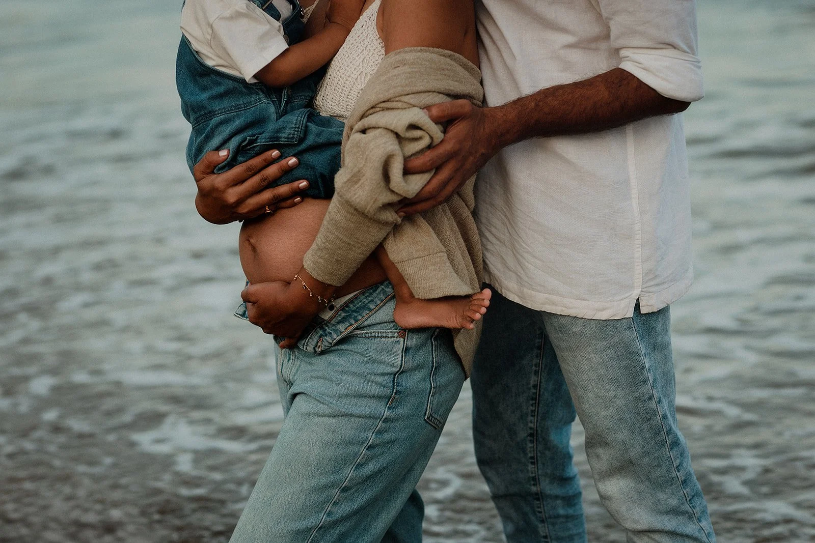 A family photograph of a husband holding on to his wife while she holds on to their Child with the sea water in the background 