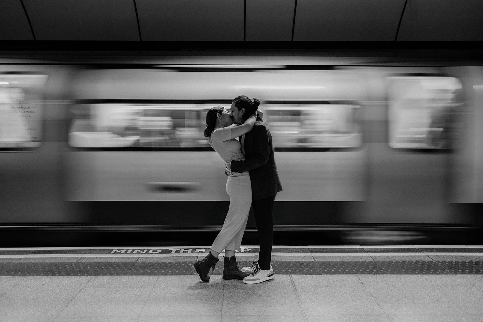 A portrait of a couple in a train station kissing as the train in the background is moving behind them 
