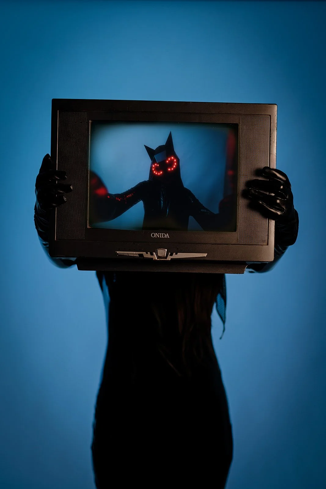 A styled portrait photograph of a person holding an old T.V. and with a mask with red lights shot by Darius N Fernandes